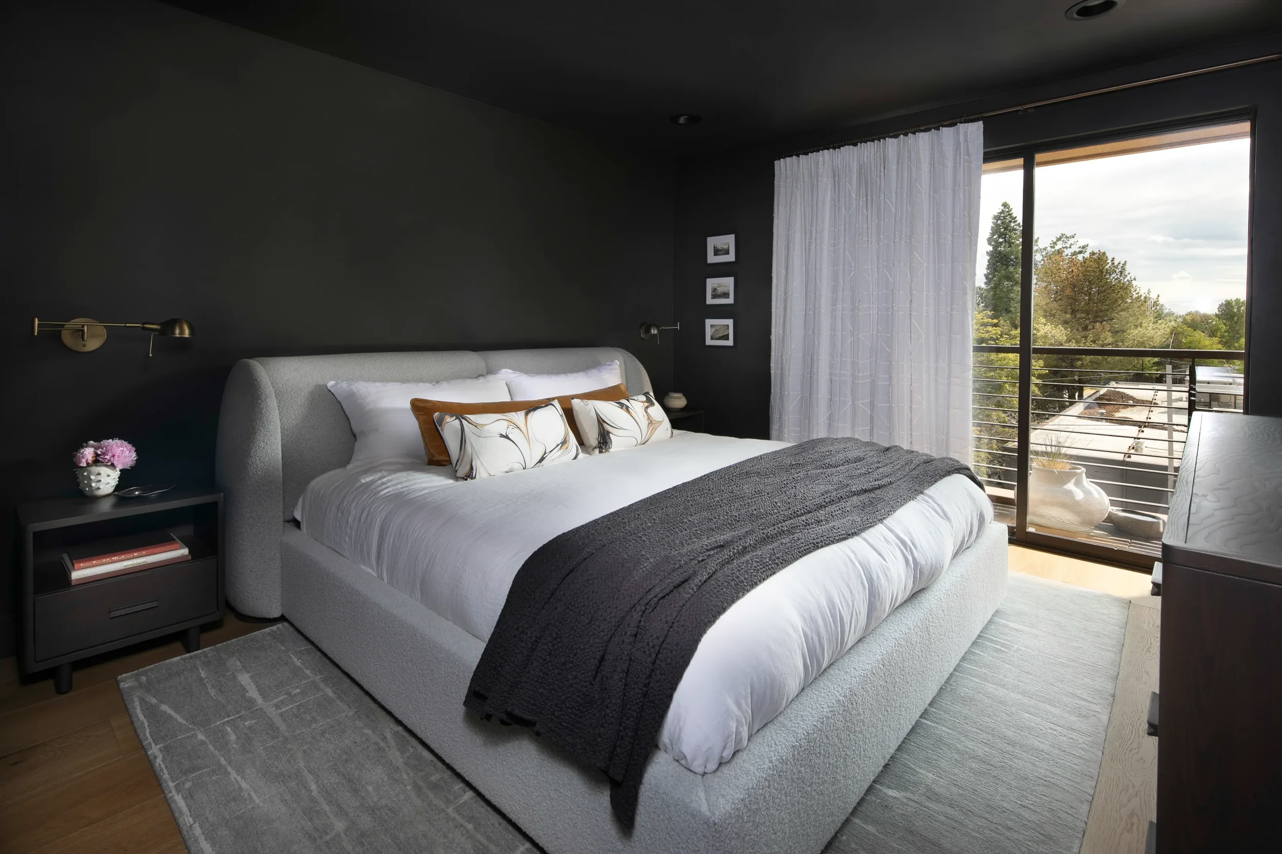Modern dark bedroom with a gray upholstered bed, white bedding, charcoal throw, bedside table, and balcony windows