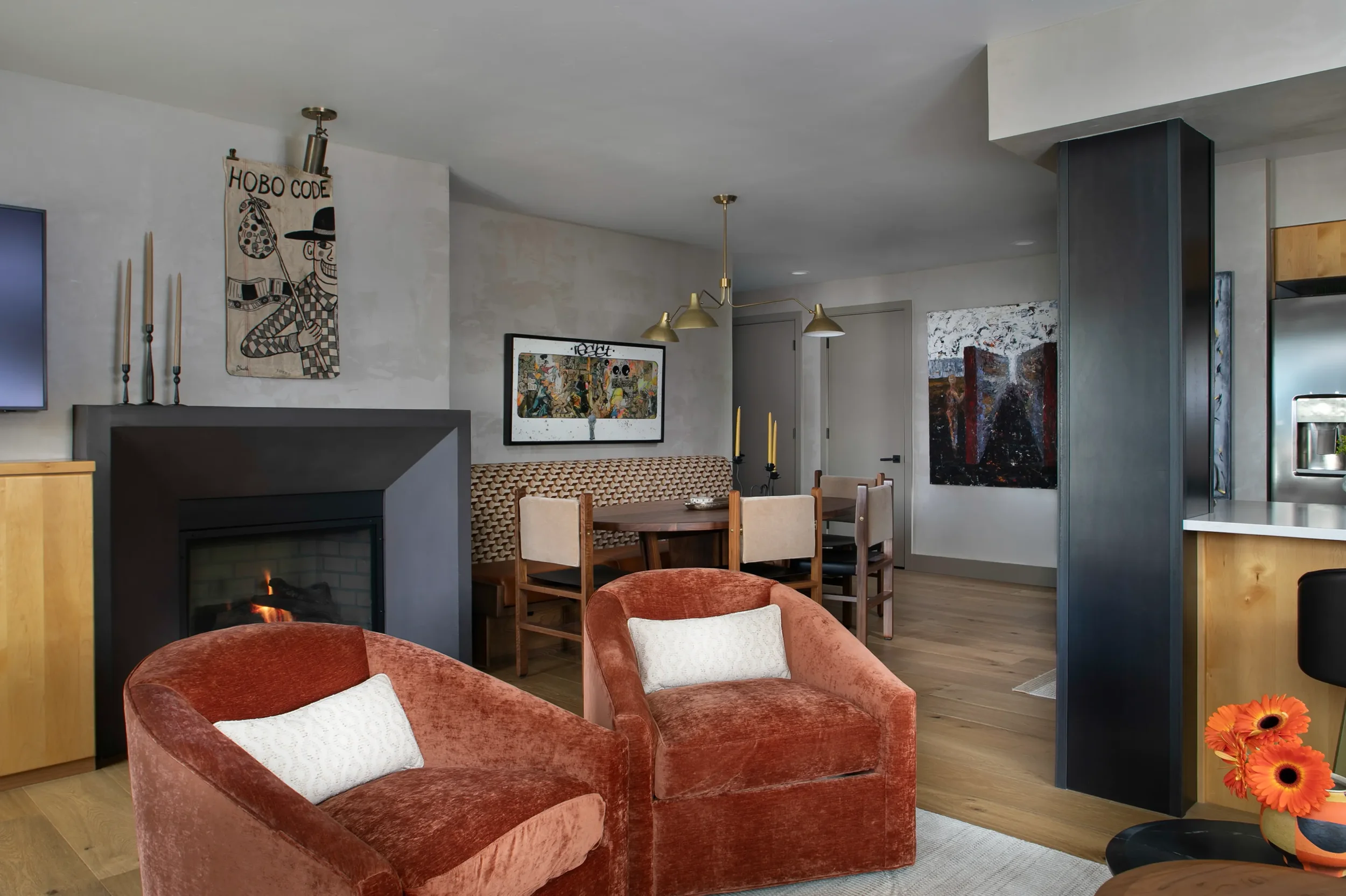 Modern open-plan living room with fireplace, rust-colored armchairs, dining table, artwork, brass pendant lights, and adjacent kitchen.