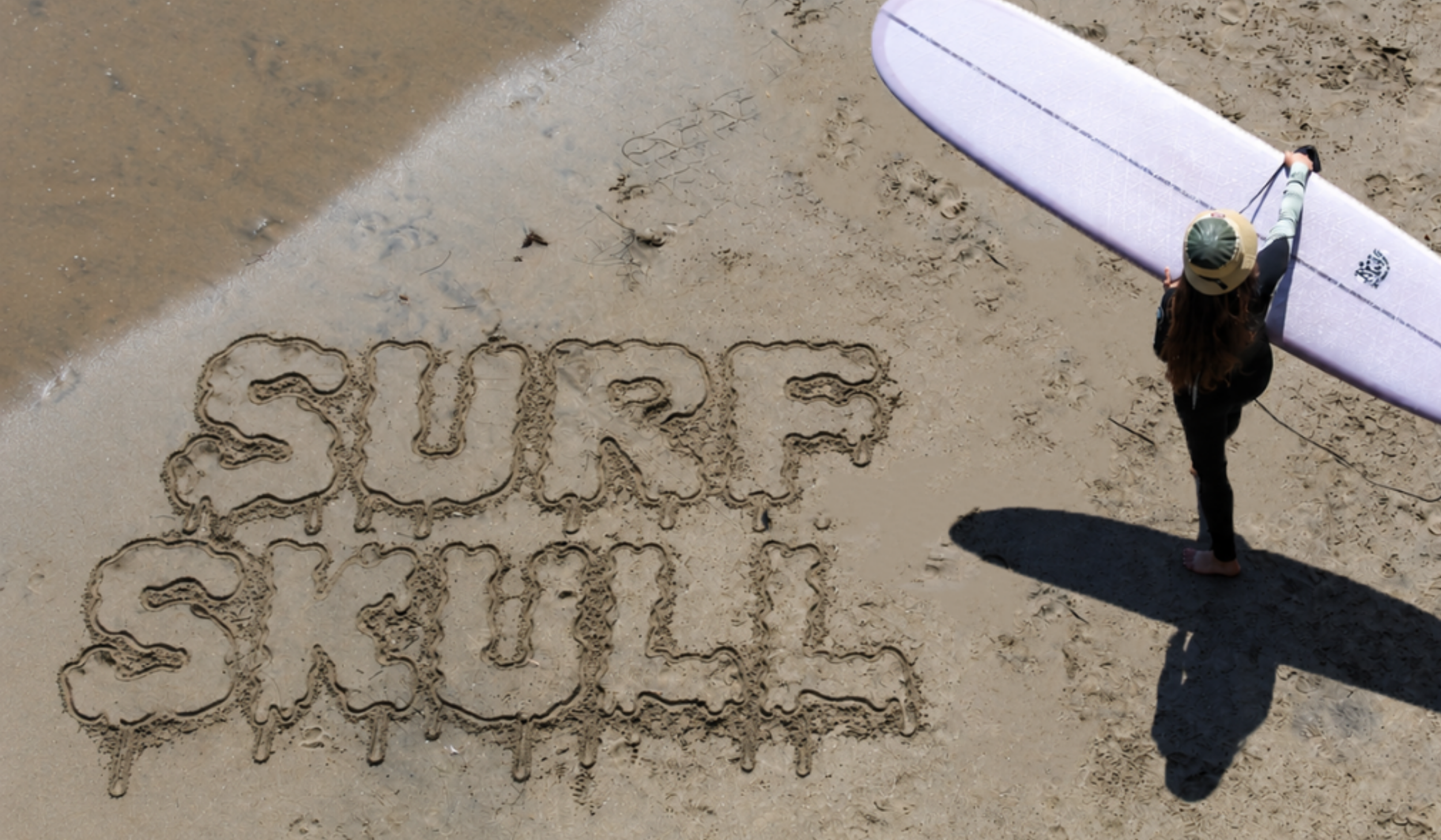 Person standing on the beach holding a paddleboard, with the words "SURF" and "SAND" written in the sand with seashells and sticks.