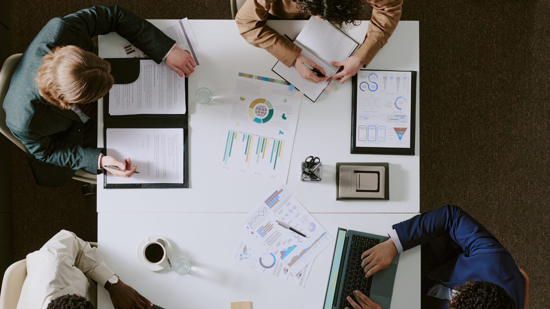 Overhead view of a business meeting with five people at a white table, discussing charts and documents. Items include laptops, papers, charts, pens, glasses of water, a coffee cup, and office supplies.