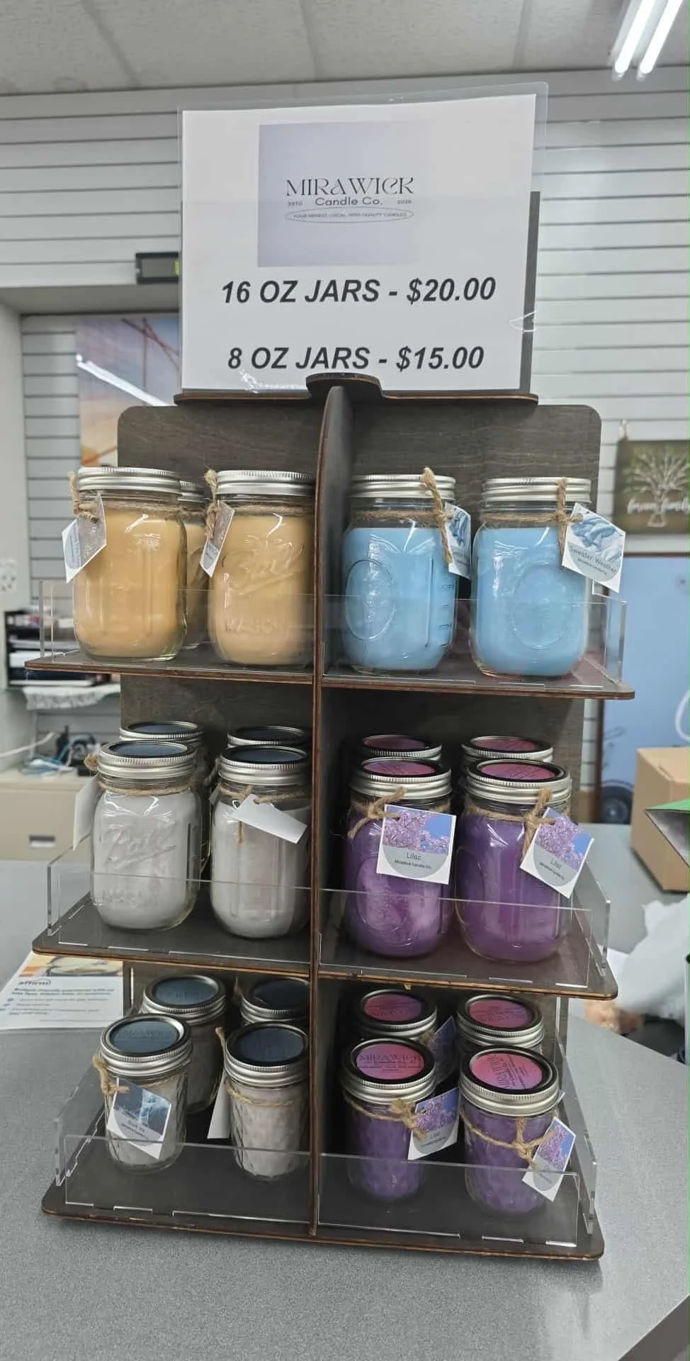 Display of scented candles in jars on a wooden shelf with signs indicating prices for 16 oz jars at $20.00 and 8 oz jars at $15.00.