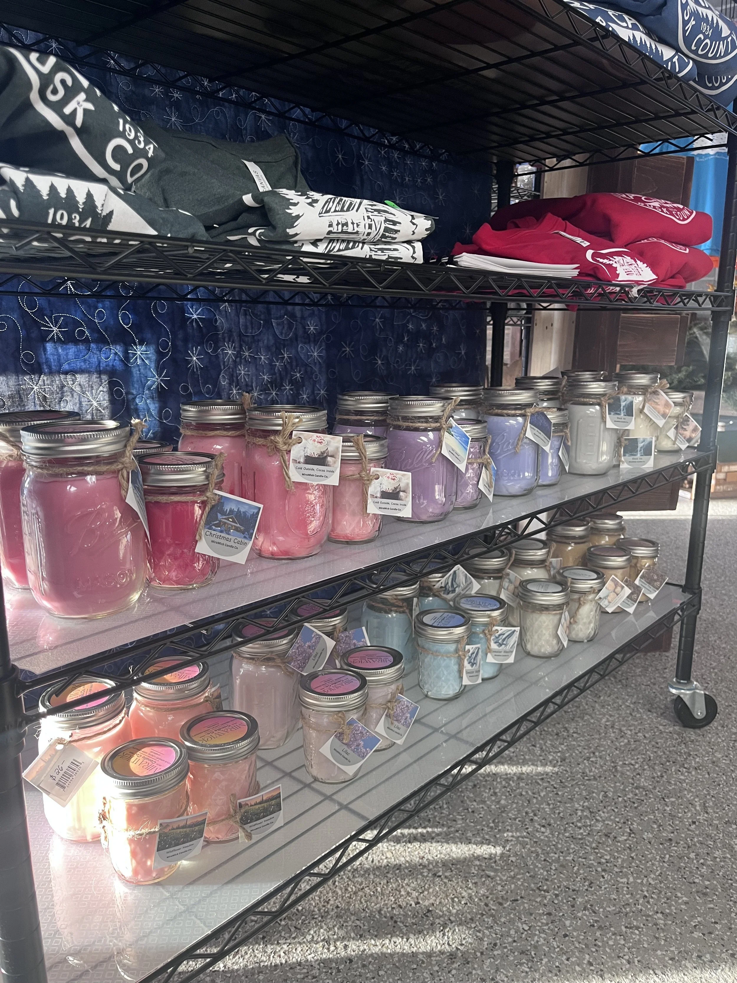 Metal shelving unit displaying candles in glass jars and folded clothing items, with a dark blue backdrop and partially visible sign.