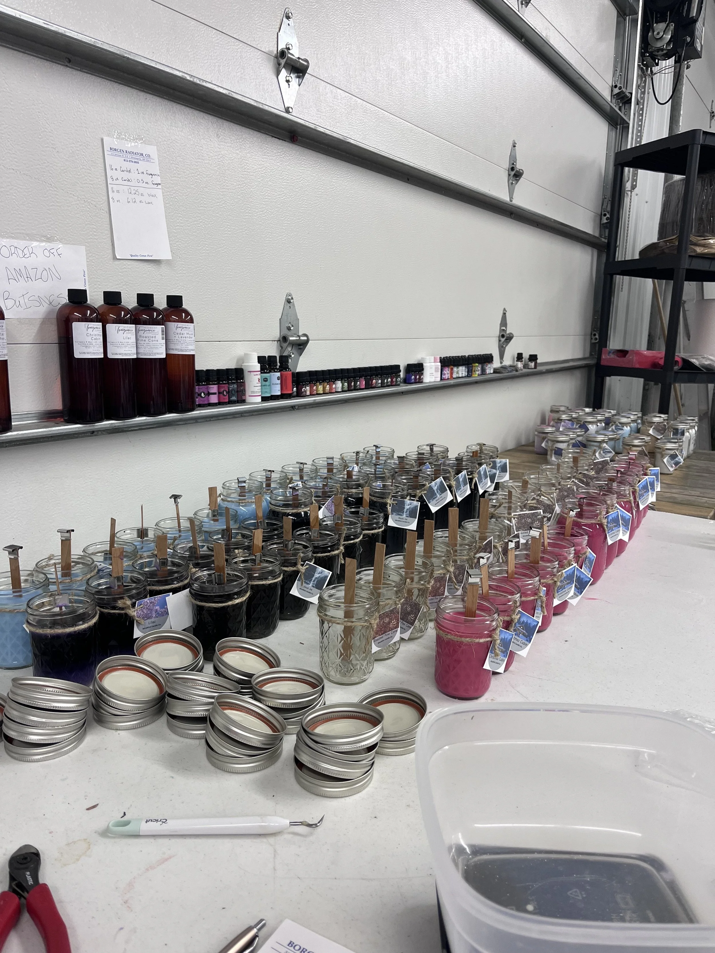 A variety of scented candles in glass jars with metal lids, arranged on a white table, with small mason jars and essential oils on a shelf behind them in a workshop or store setting.