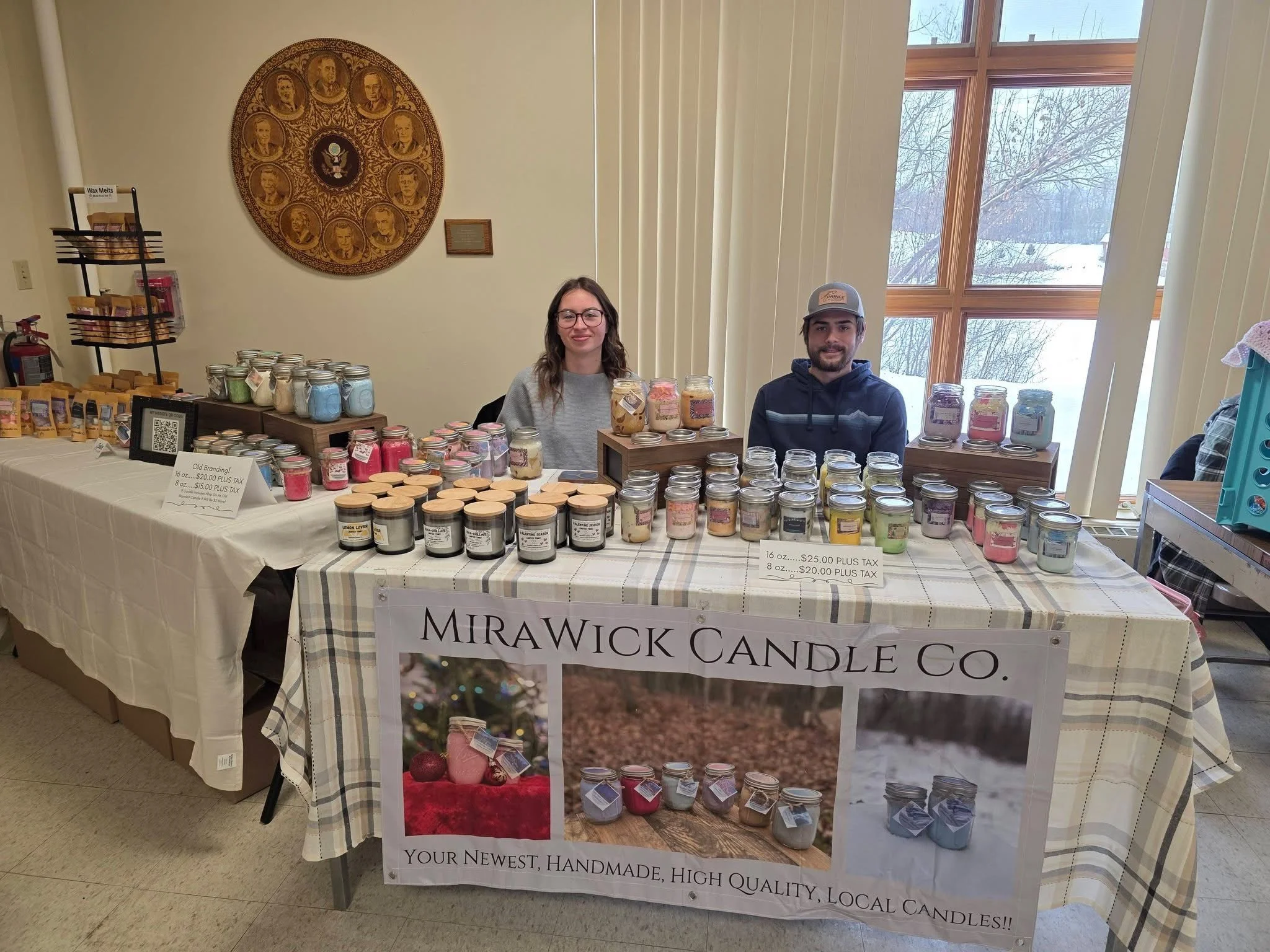 Two people sitting behind a table display filled with handmade candles at a craft fair. A banner reads "Mirawick Candle Co. Your newest, handmade, high quality, local candles!" The setting appears to be indoors with large windows showing snowy trees outside.