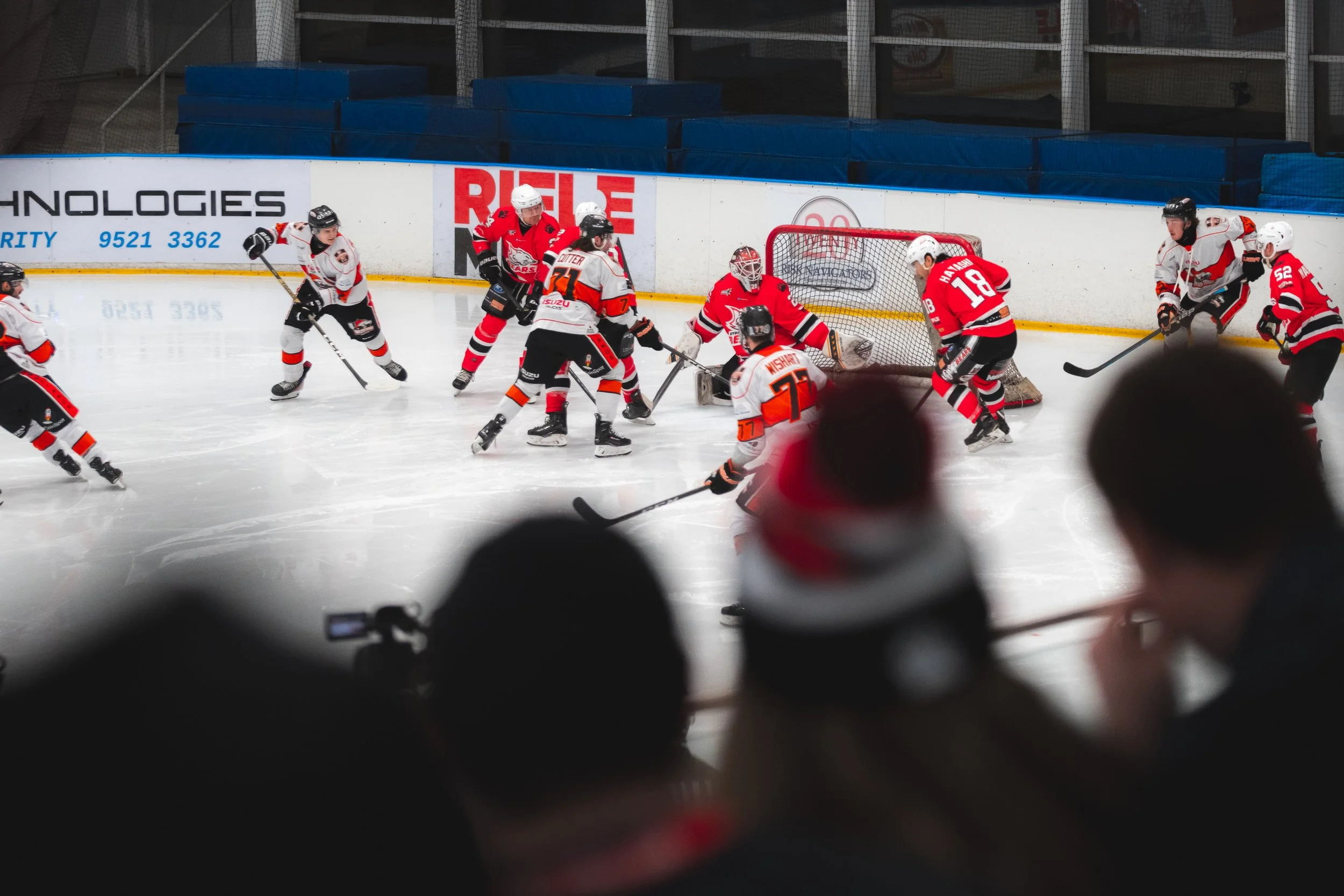 An ice hockey game with players from two teams in red and white uniforms battling near the goal. Spectators are watching from the stands.