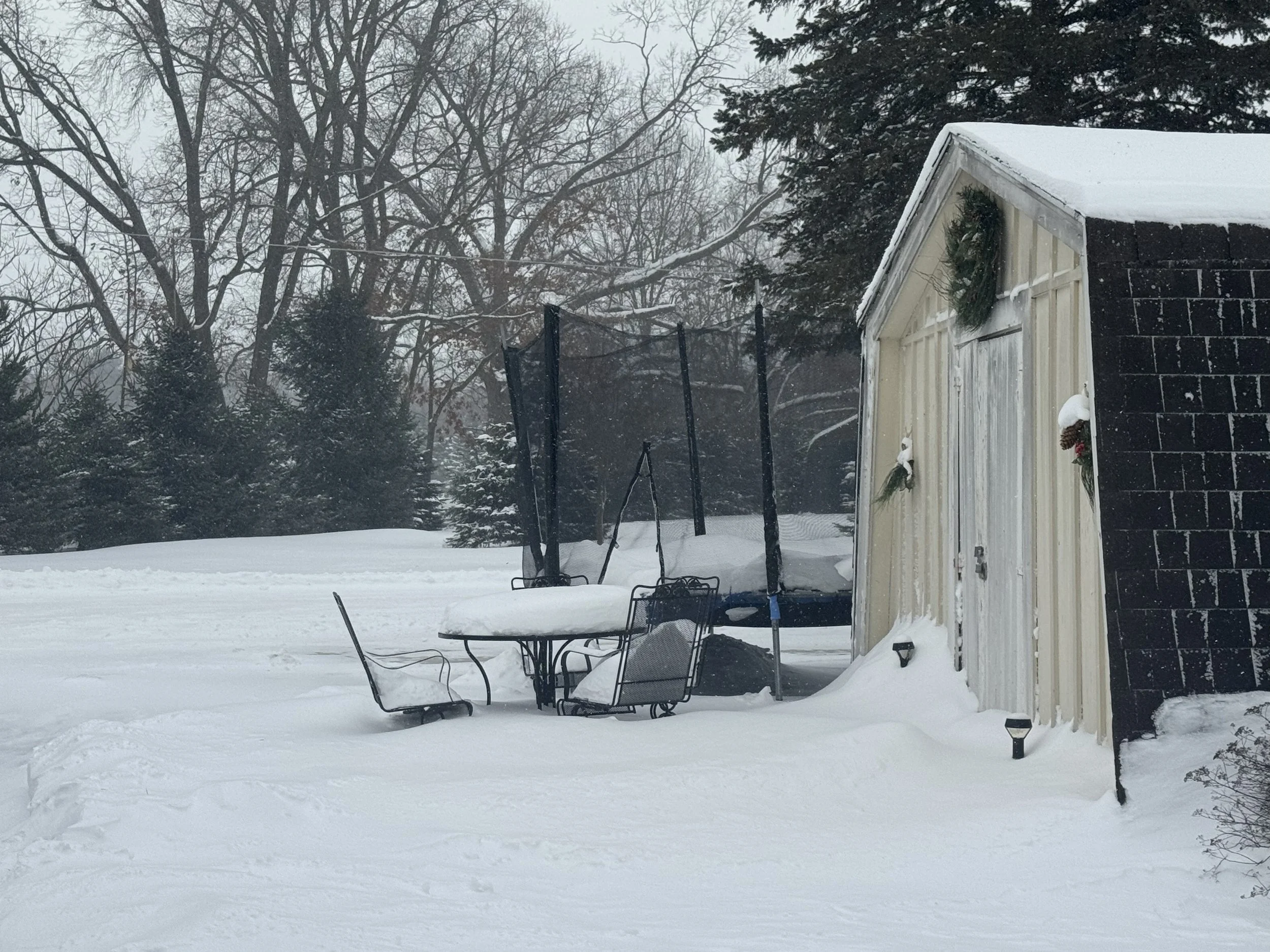 Snow-covered backyard with outdoor table and chairs, a trampoline, and a shed decorated with holiday wreaths, surrounded by leafless trees in winter.