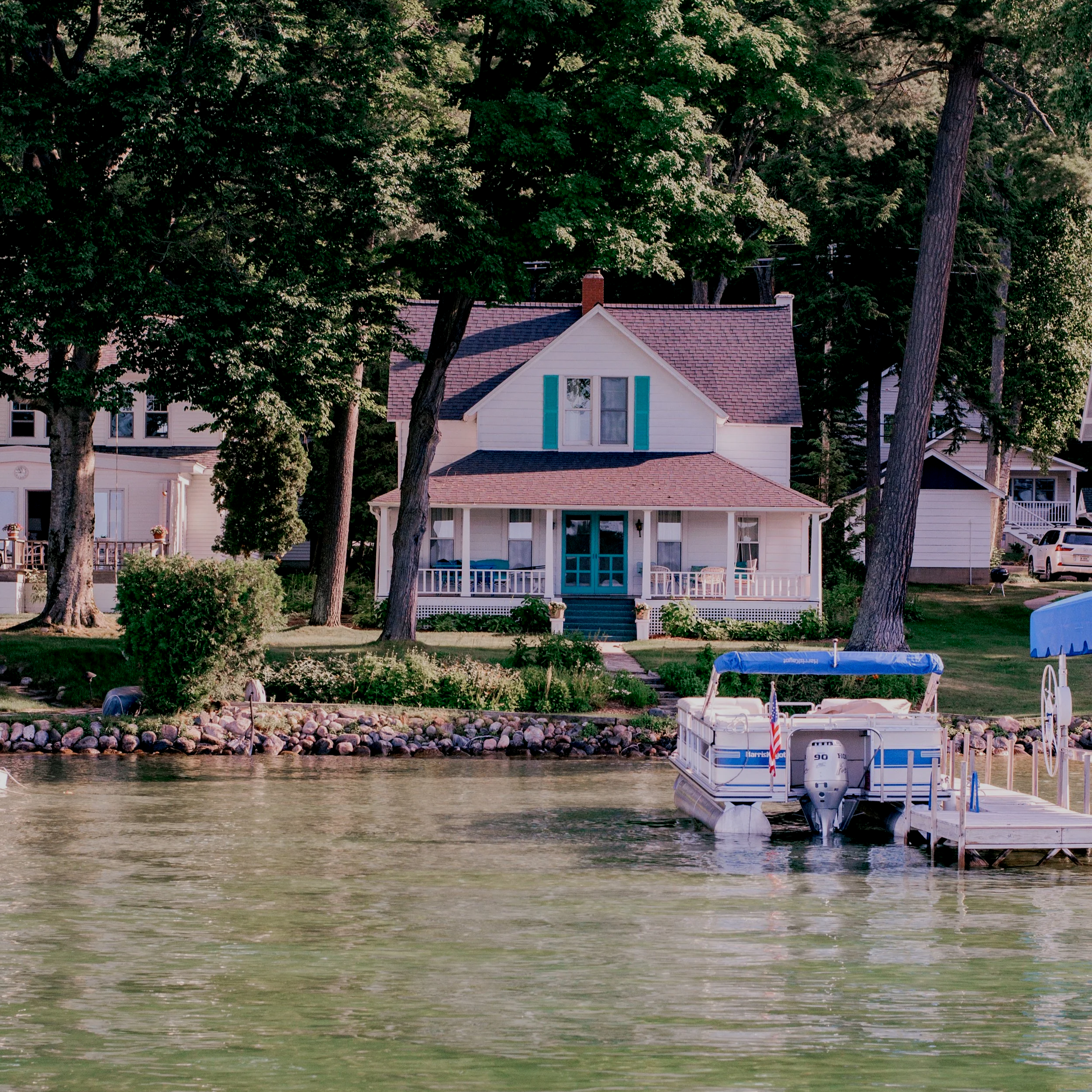 A house with a porch facing a lake, with a boat docked nearby. The house is surrounded by tall trees and has a well-kept lawn with decorative bushes.