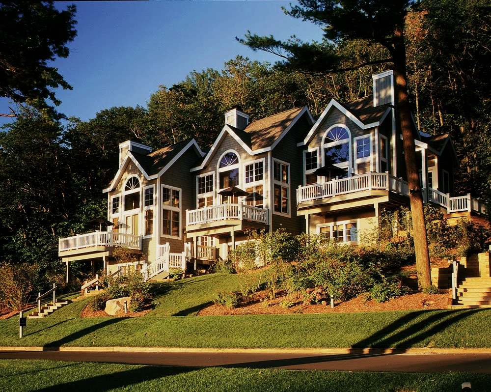 Three multi-story houses with large windows, balconies, and peaked roofs, situated on a hillside with landscaped greenery and trees in the background.