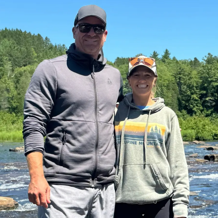 A man and woman standing outdoors near a river with a lush green forest in the background, smiling at the camera during daytime.