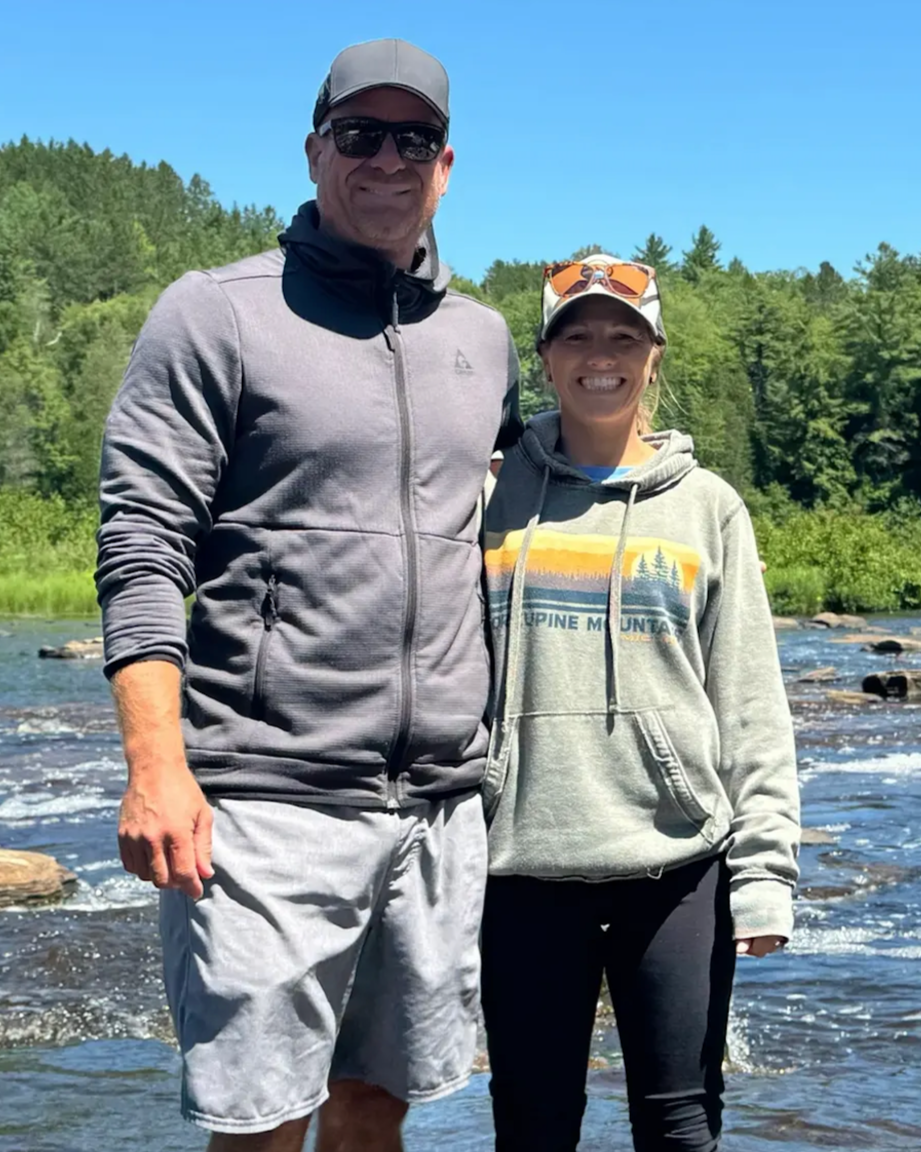 A man and woman standing side by side outdoors by a river, smiling, surrounded by trees and clear blue sky, dressed in casual outdoor clothing.