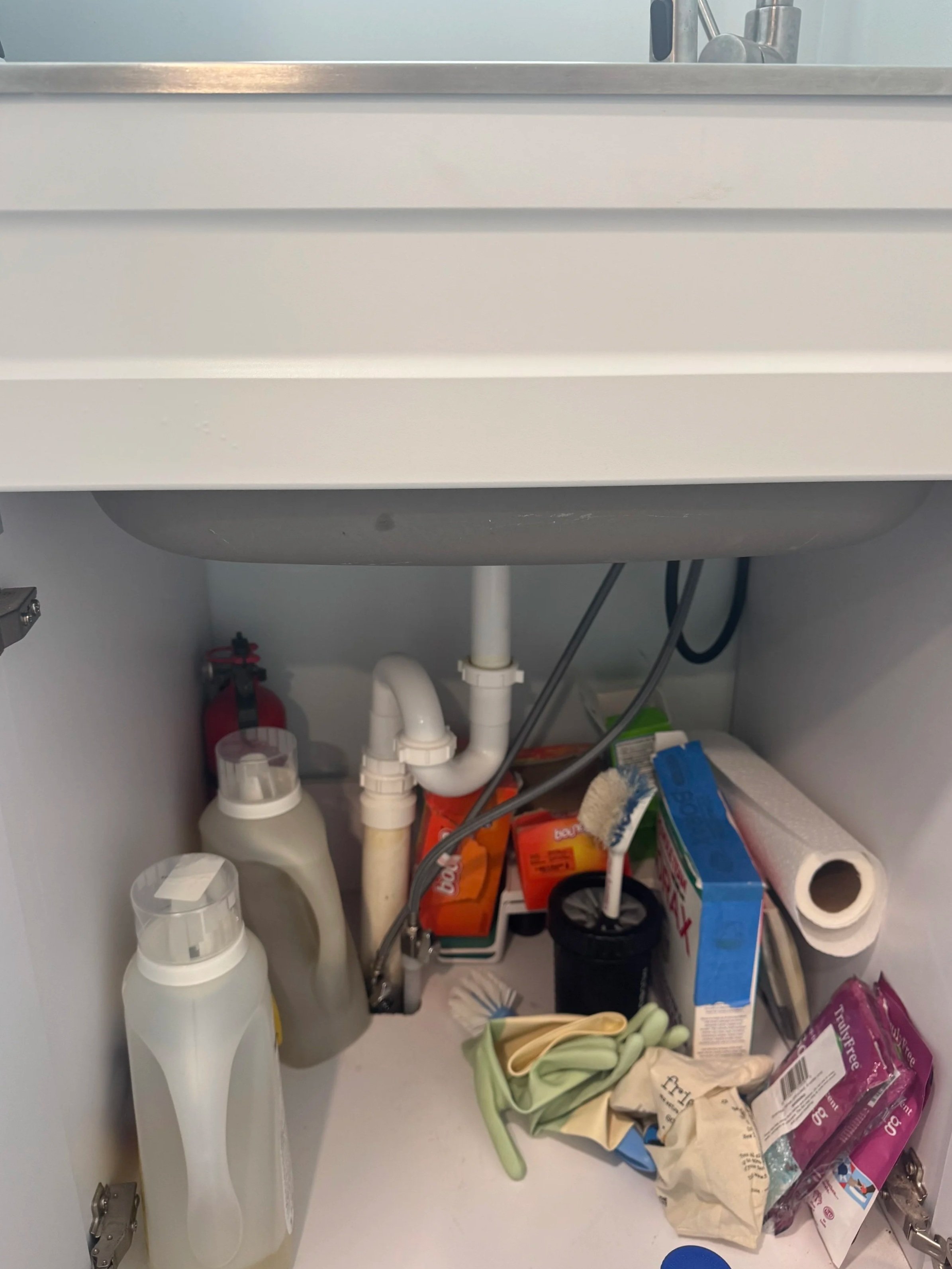 Open kitchen cabinet beneath the sink with cleaning supplies, paper towels, and miscellaneous items inside.