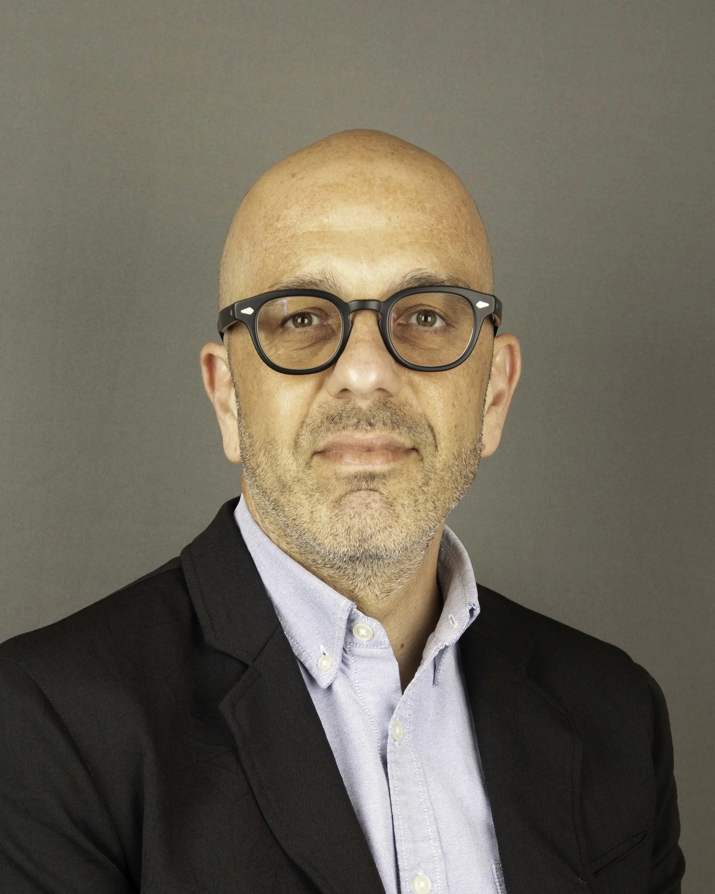 A man wearing glasses, a light blue dress shirt, and a black suit jacket, with a bald head and some facial hair, looking directly at the camera.