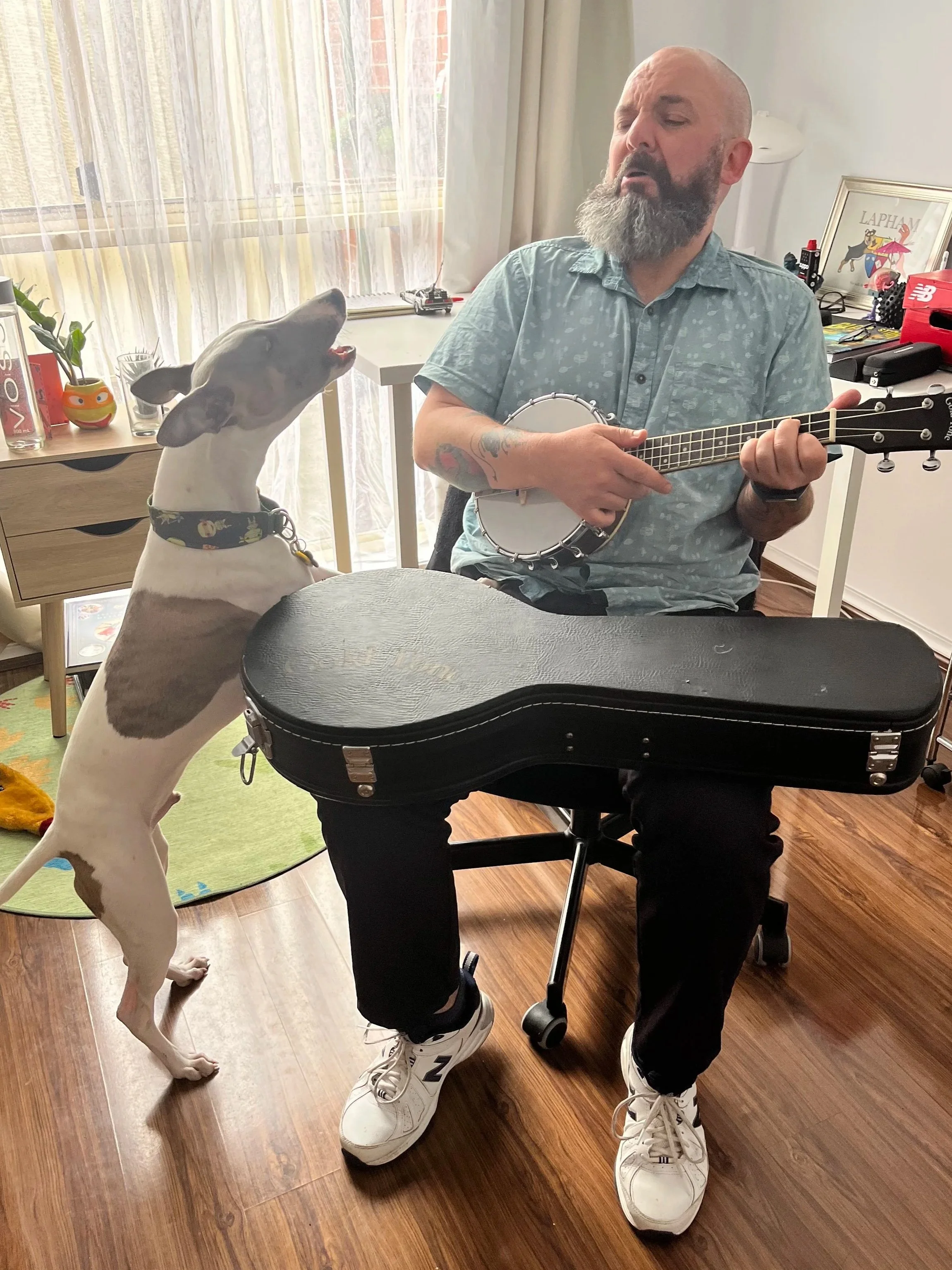Fabian sitting in a chair, playing a banjo-ukulele and singing along with a whippet that has rested its front paws on his leg as it howls.