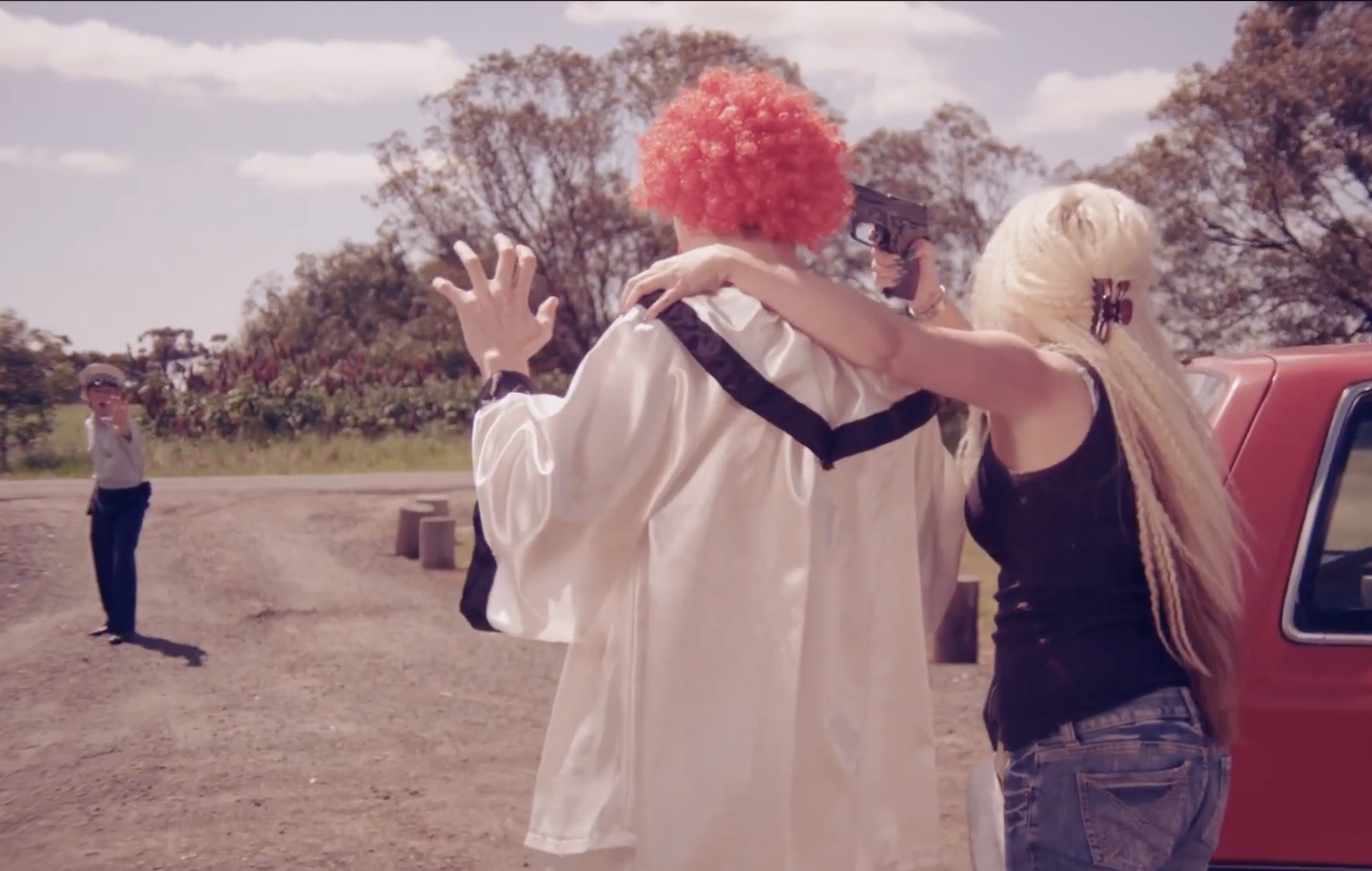 A woman in a rural carpark holds a gun to a clown's head. The clown has his hands in the air in surrender. Their backs are to us. In the distance, a police officer approaches them, holding out their hand in a calming manner.