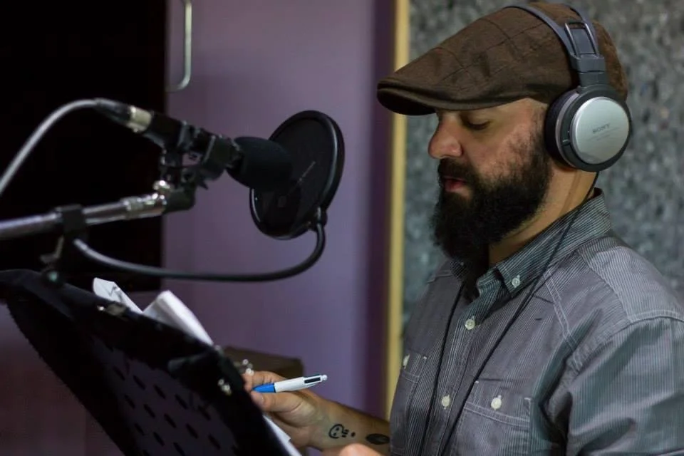 Fabian Beevers-Lapham in a recording studio. He is wearing headphones, a grey shirt, and a brown cap. He has a beard. He is holding a script and a pen, and standing next to a microphone stand that holds a mic and a pop filter.
