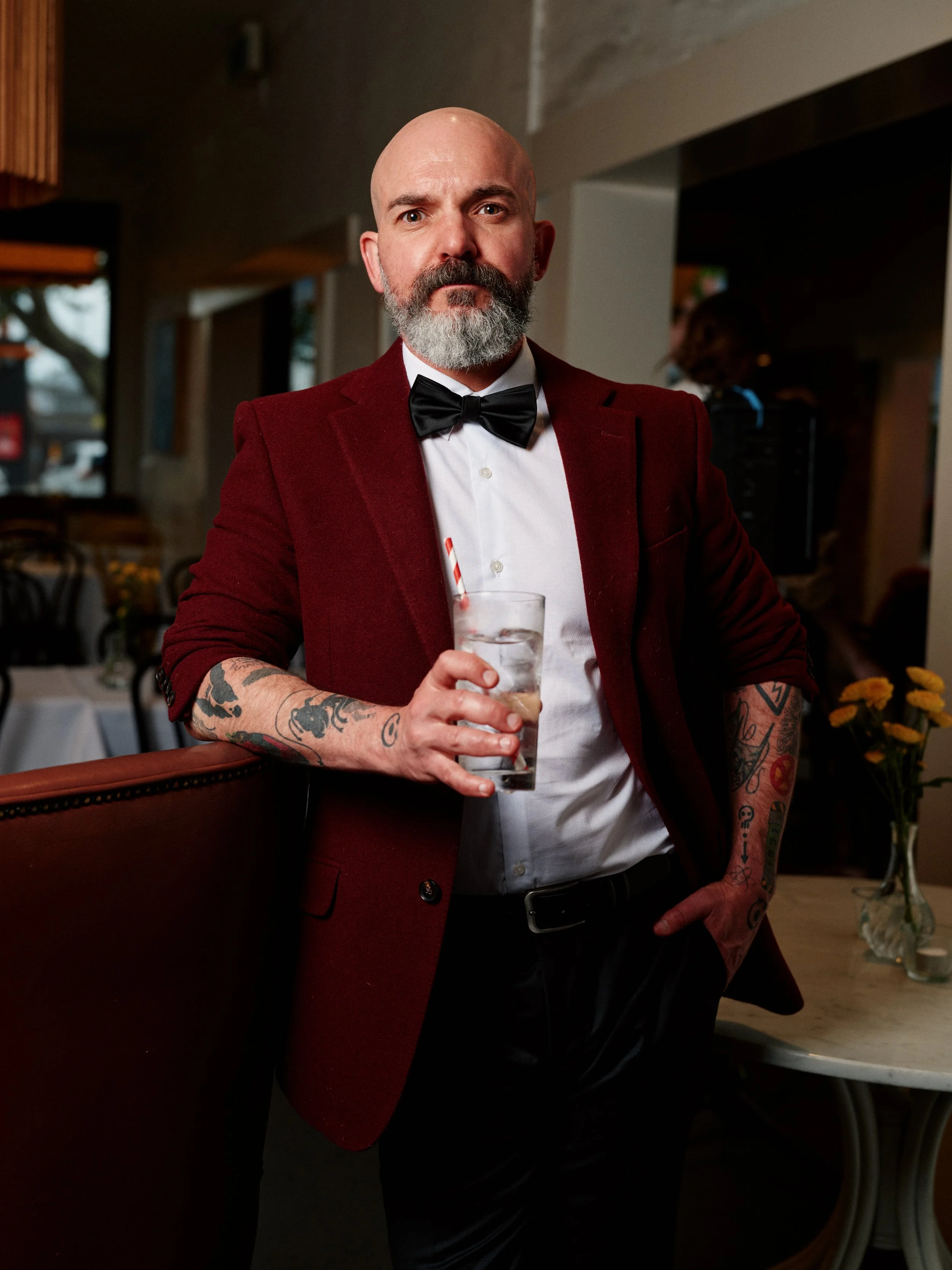 Fabian Beevers-Lapham wearing a burgundy suit jacket, white shirt, and a black bowtie. He is holding a glass of ice water. He has rolled up sleeves, tattoos on his forearms, a beard and a shaved head.