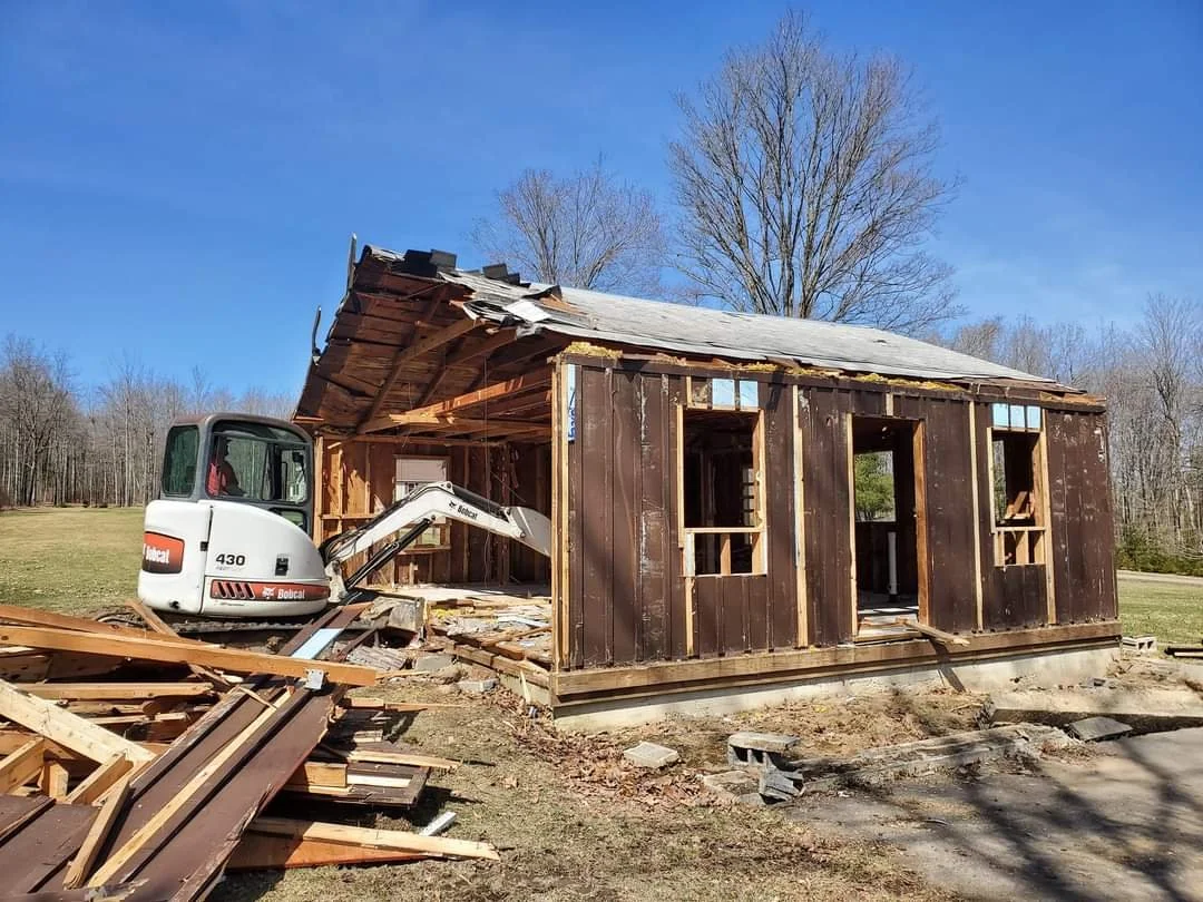 A small wooden house being demolished with an excavator in a grassy area on a clear day.