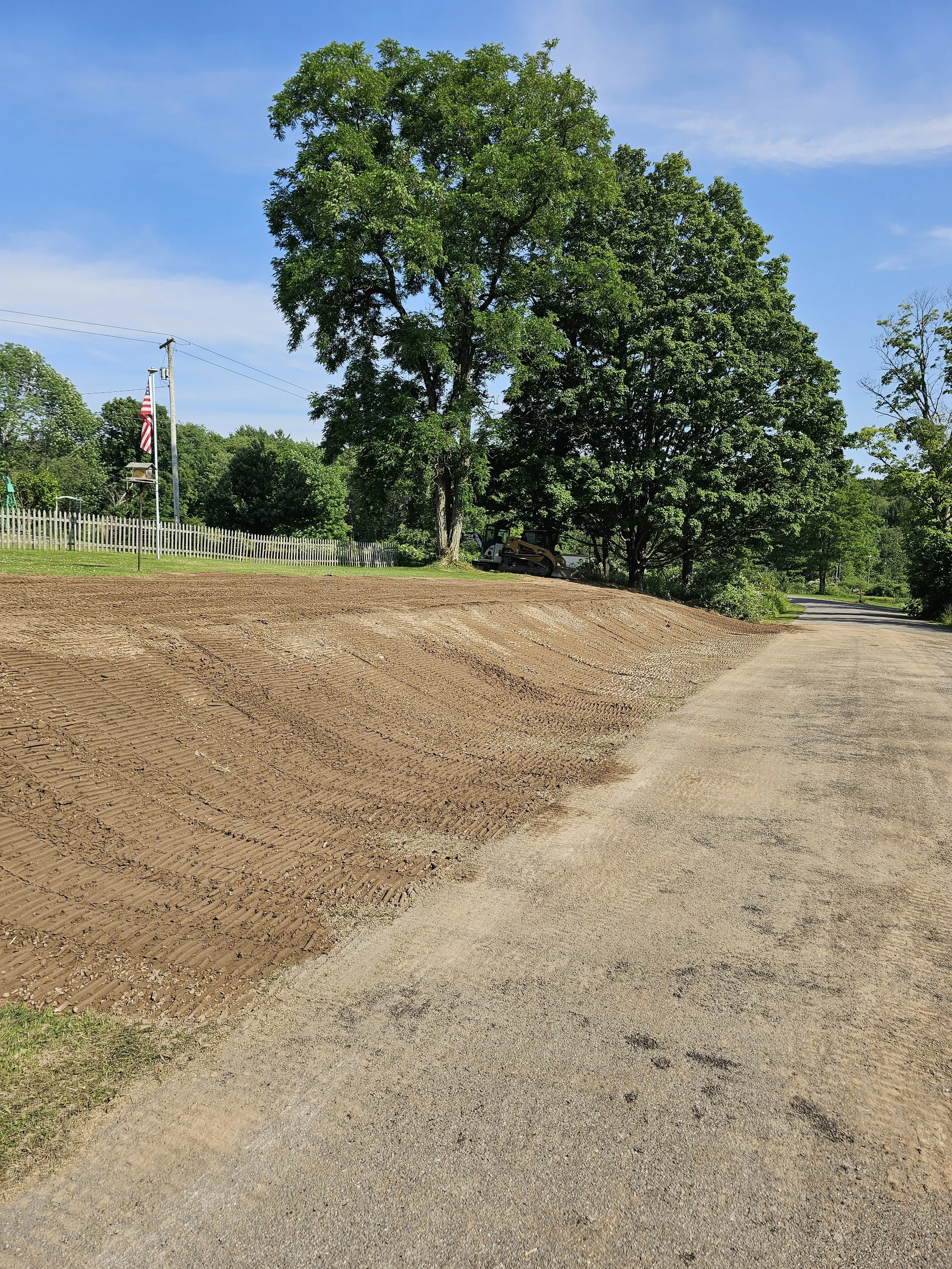 A dirt road next to a freshly tilled garden bed bordered by trees, with a blue sky and a few clouds in the background.