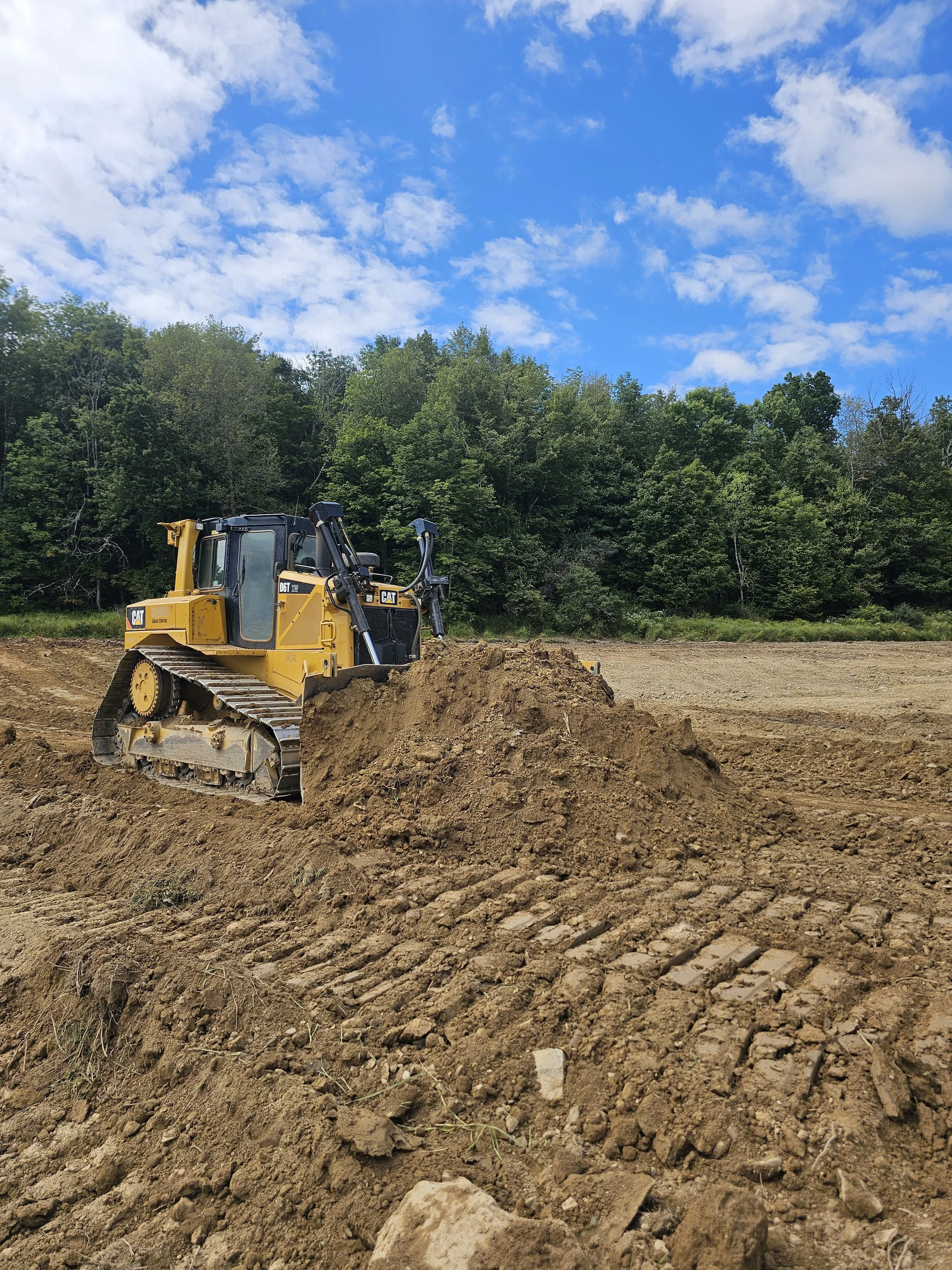 A yellow Caterpillar bulldozer pushing soil on a construction site with green trees and a partly cloudy sky in the background.