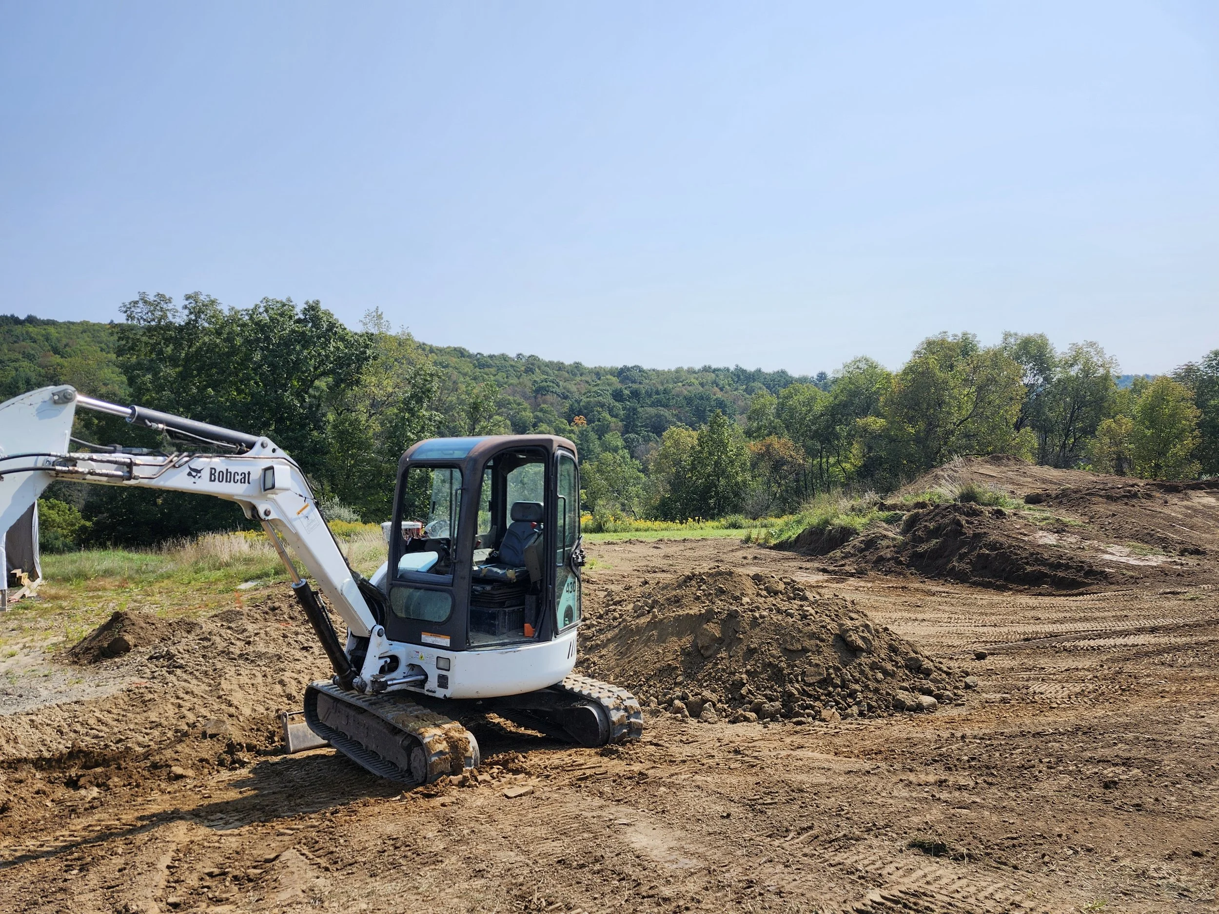 Construction site with a small excavator and dirt mounds, surrounded by green trees under a clear blue sky.