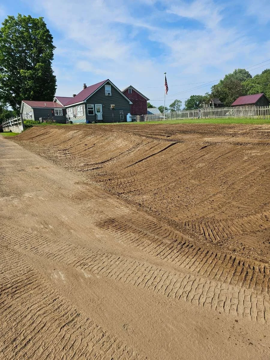 A residential area with a house, American flag, and a fenced yard. The foreground shows freshly graded dirt or construction site with tire tracks.