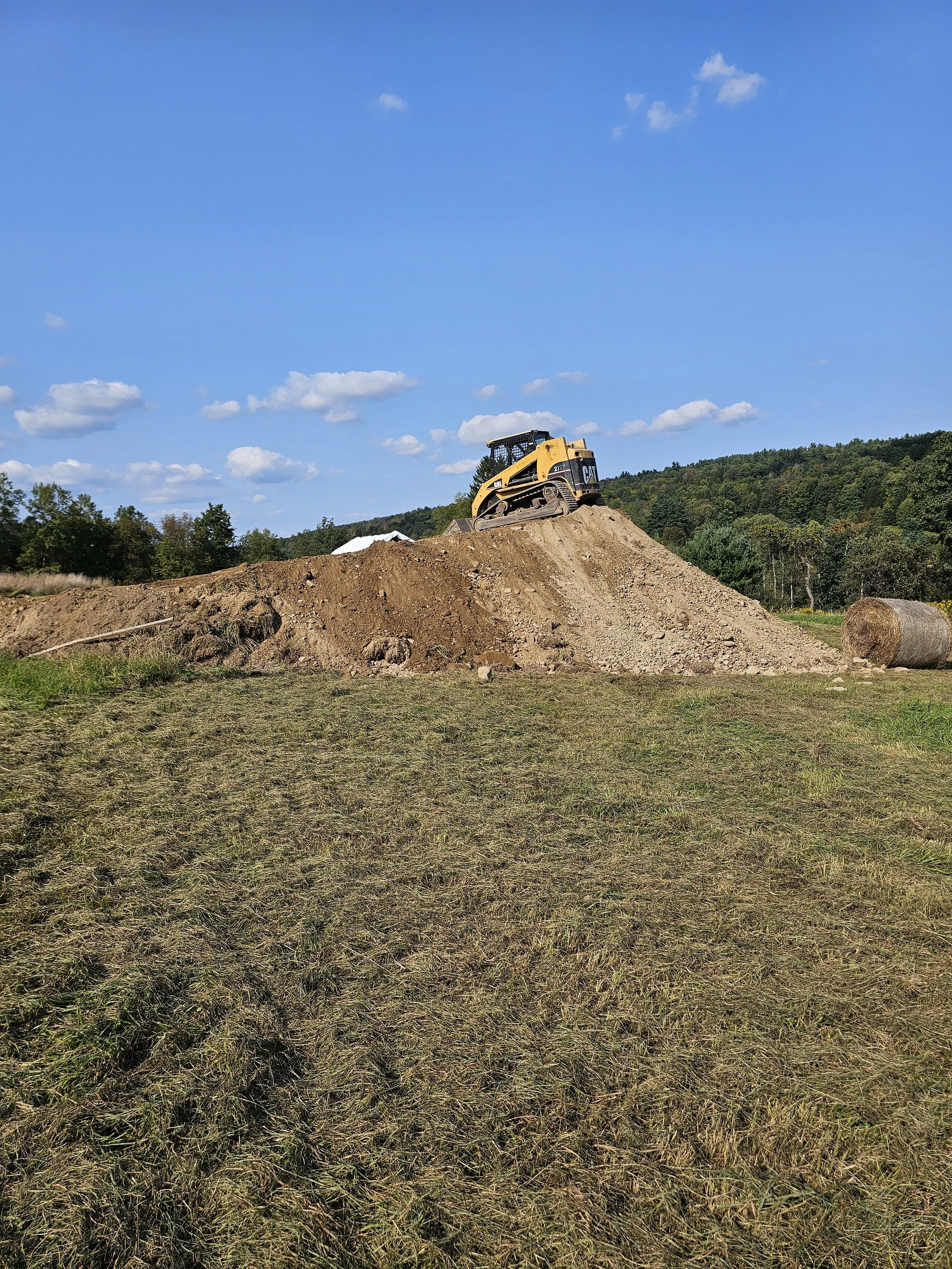 A yellow Caterpillar bulldozer on a mound of dirt, with a clear blue sky and trees in the background.