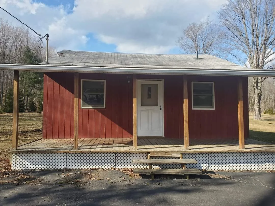 A small red house with a porch under construction, featuring wooden posts and steps, a white door, and two windows, surrounded by trees and a partly cloudy sky.