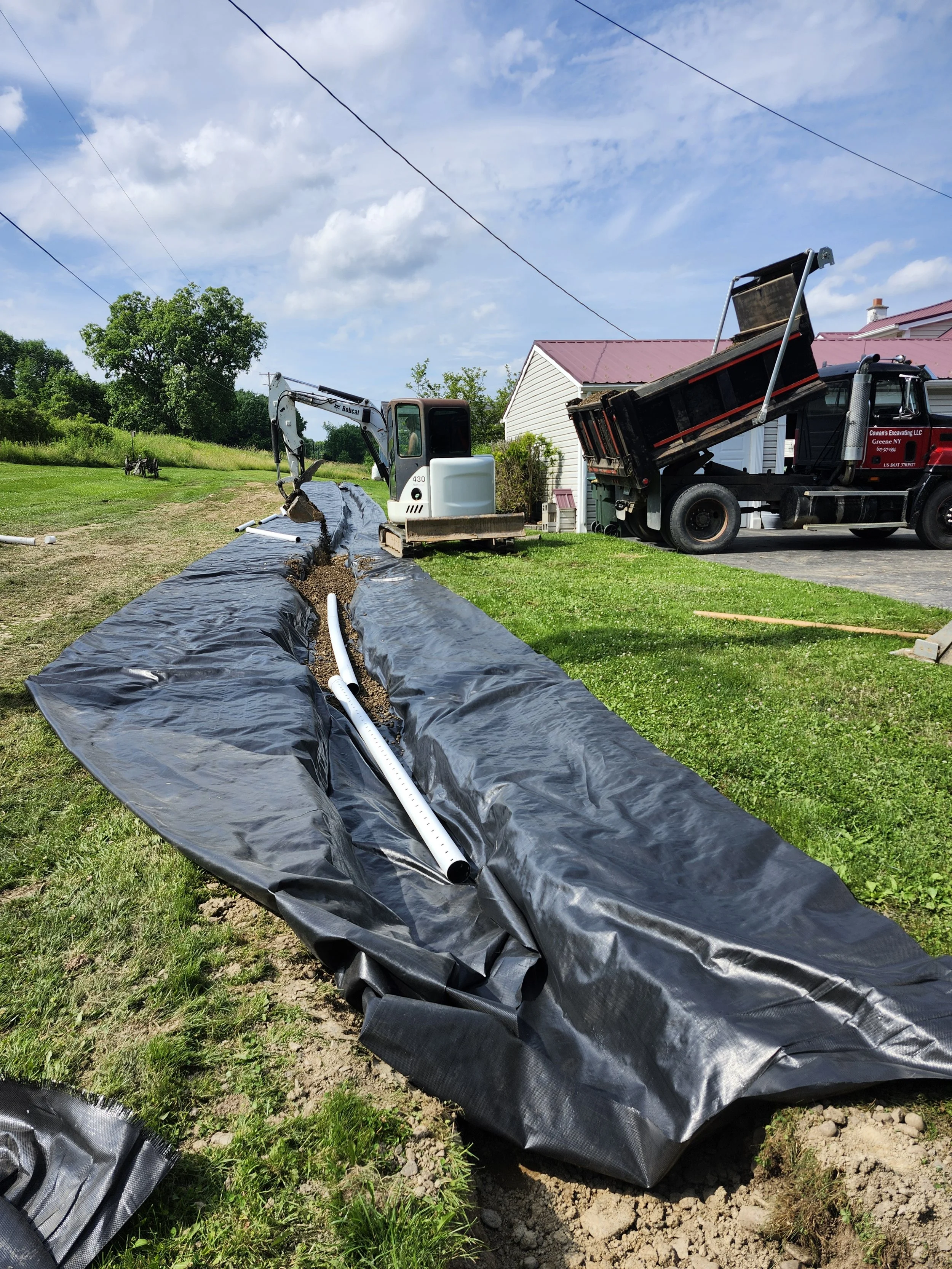 Construction site with a small excavator digging a trench lined with black plastic sheeting, pipes are being installed. A dump truck is parked nearby with its bed raised. Residential houses and green trees are visible in the background under a partly cloudy sky.