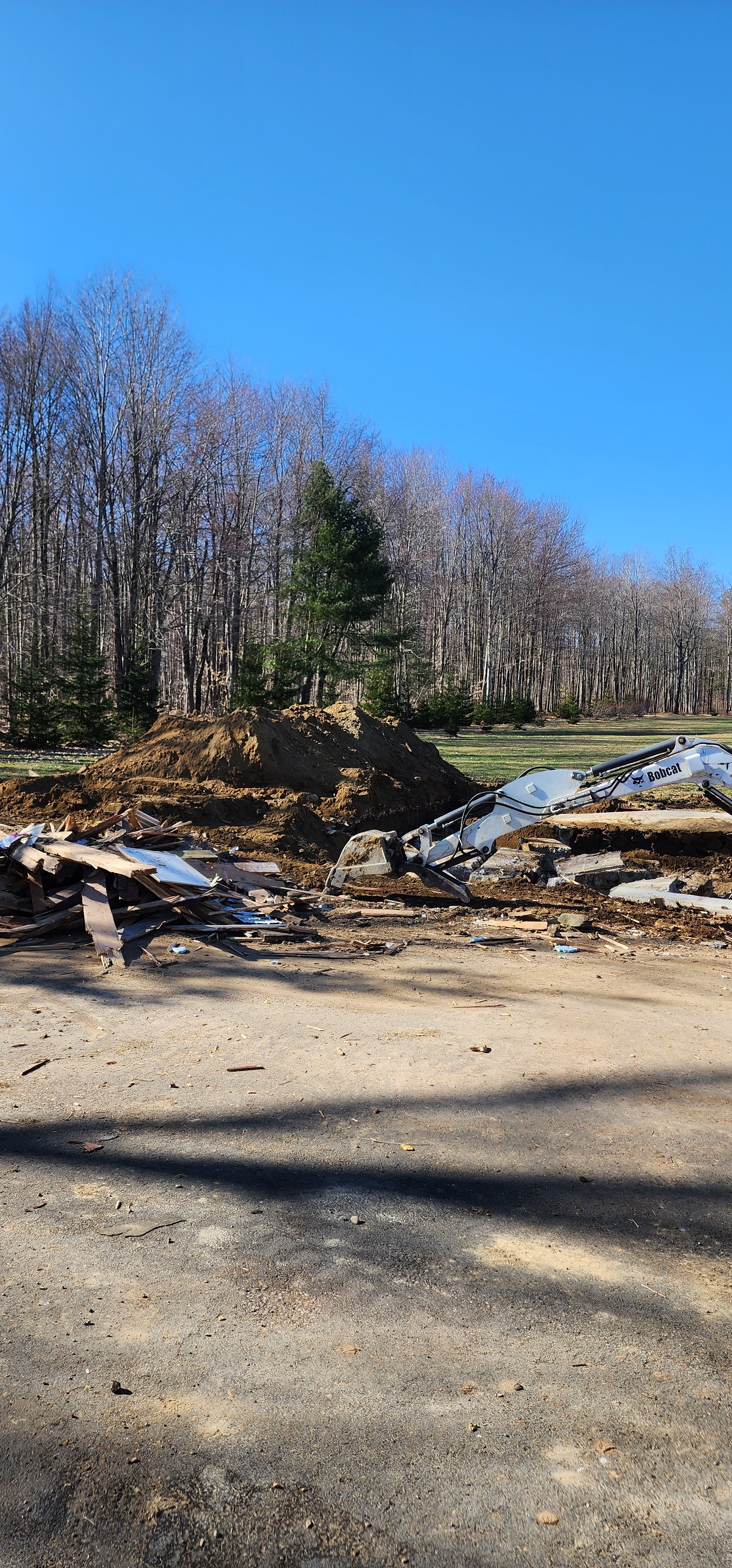 Construction site with a Bobcat excavator and a pile of dirt and debris on a gravel surface, with a background of leafless trees and a clear blue sky.