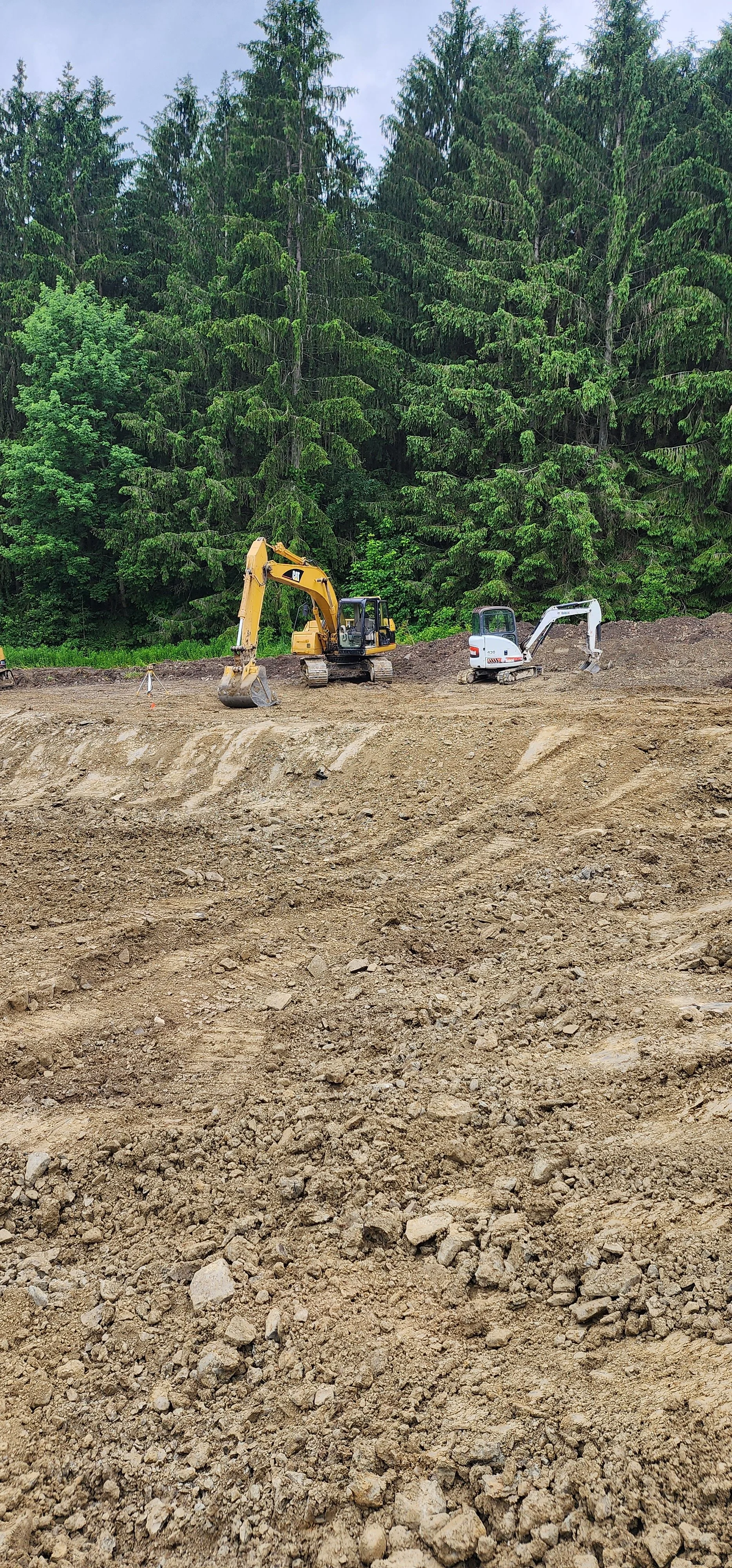 Construction site with two excavators, one yellow and one white, working on a dirt area surrounded by green trees.