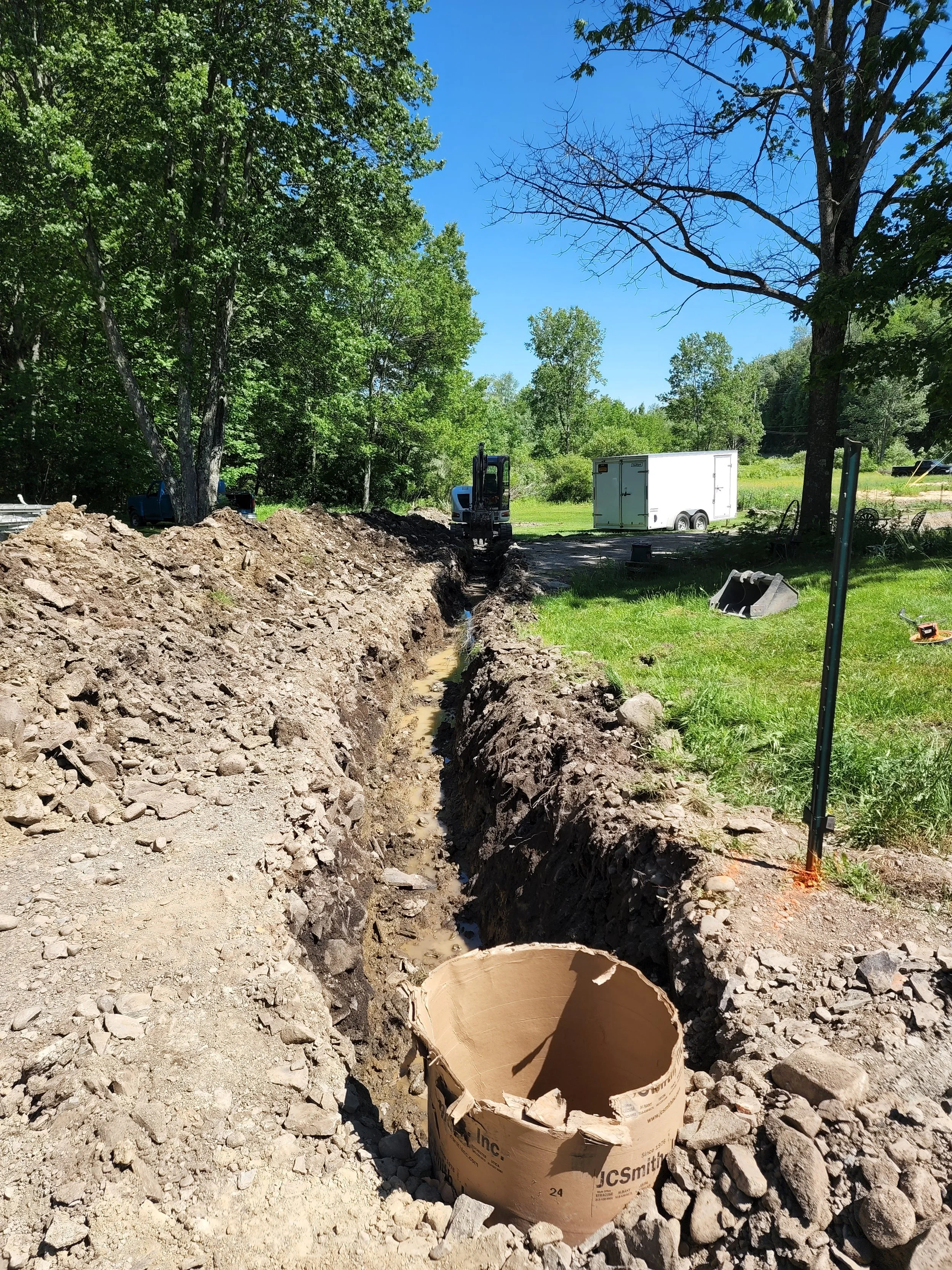 Excavation site with a narrow trench alongside a grassy area, a small excavator in the distance, a white trailer, and construction equipment, surrounded by trees on a sunny day.