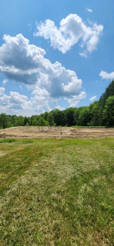 A grassy field with some trees in the background under a partly cloudy sky, and a small construction or excavation site in the middle distance.