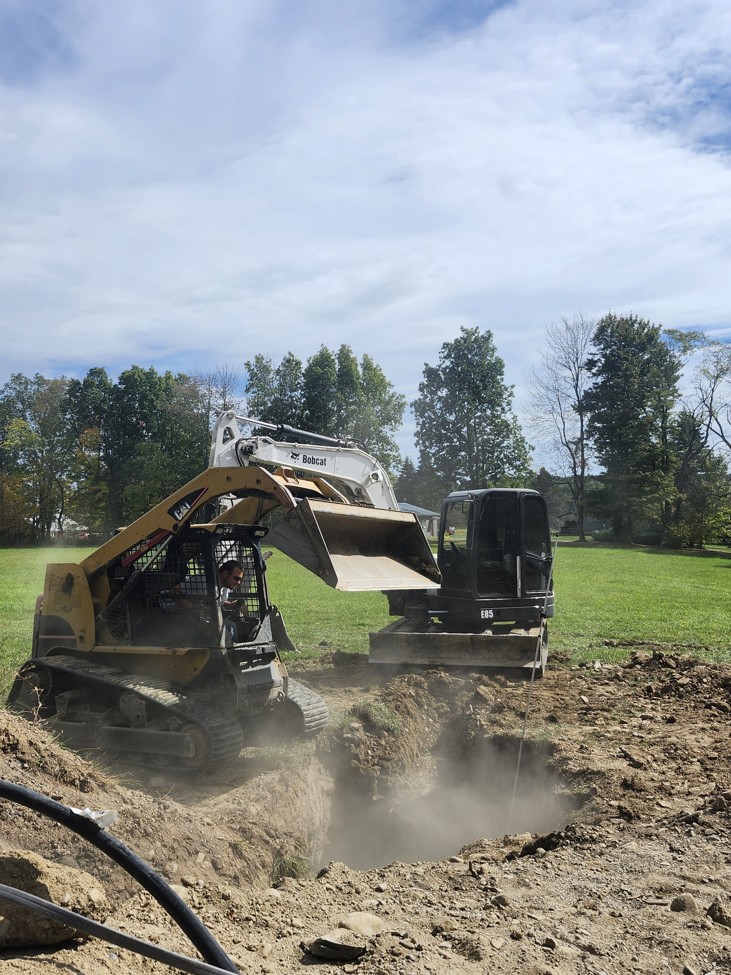 Construction workers operating a small excavator and a Bobcat machine digging a hole in the ground on a grassy field with trees in the background and a partly cloudy sky.