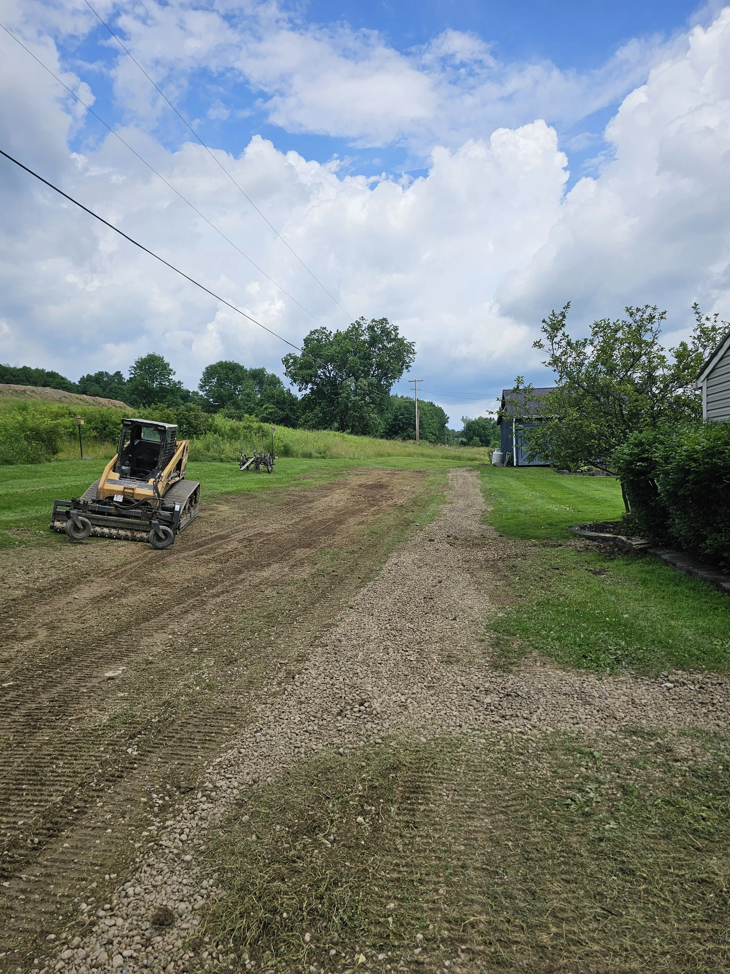 A gravel and dirt pathway in a rural yard with a skid-steer loader on the left and a small blue shed on the right, trees and a partly cloudy sky in the background.