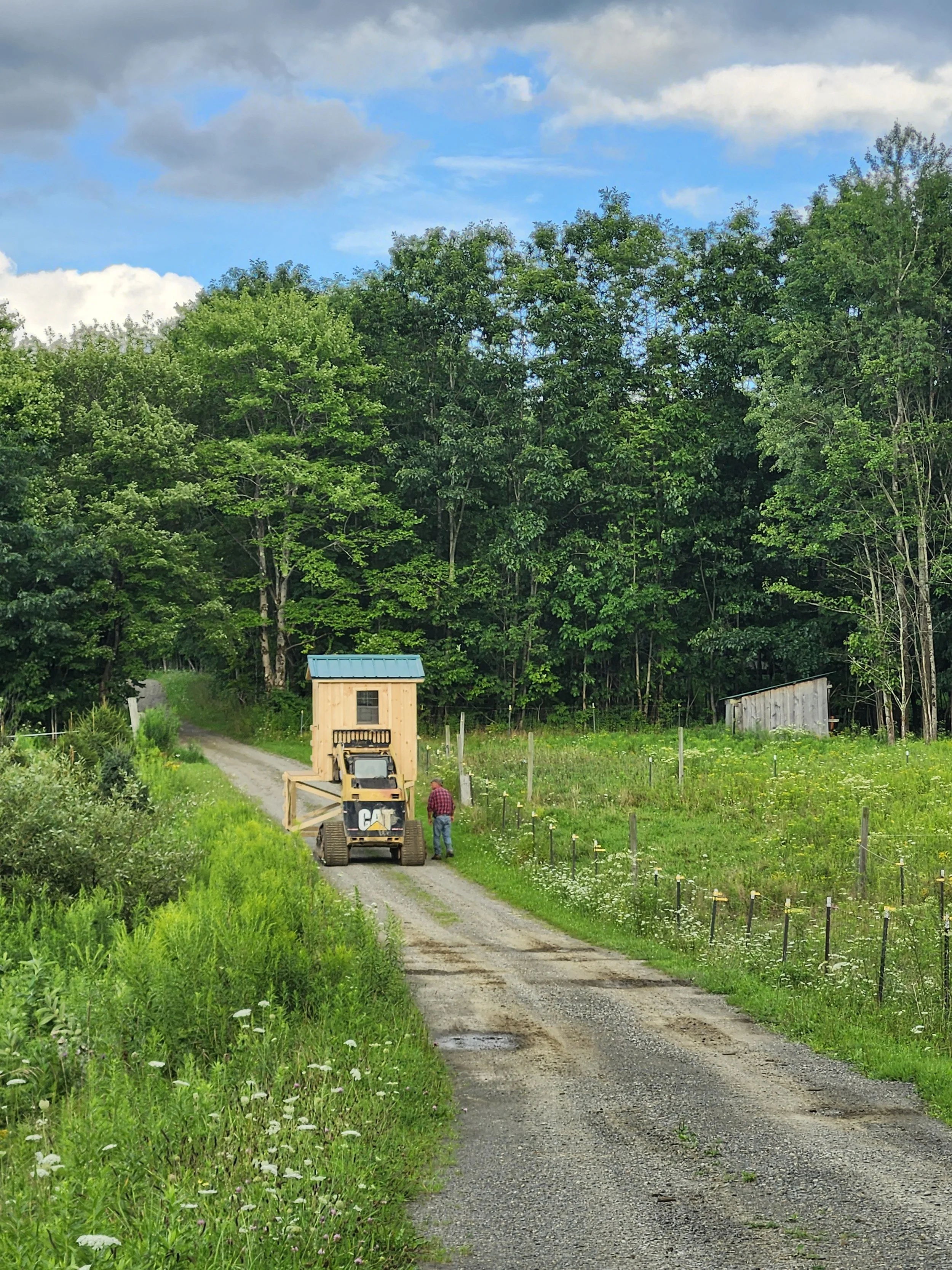 A rural dirt road with a man standing next to a small construction vehicle on a grassy hillside. There are green trees in the background and a partly cloudy blue sky above.