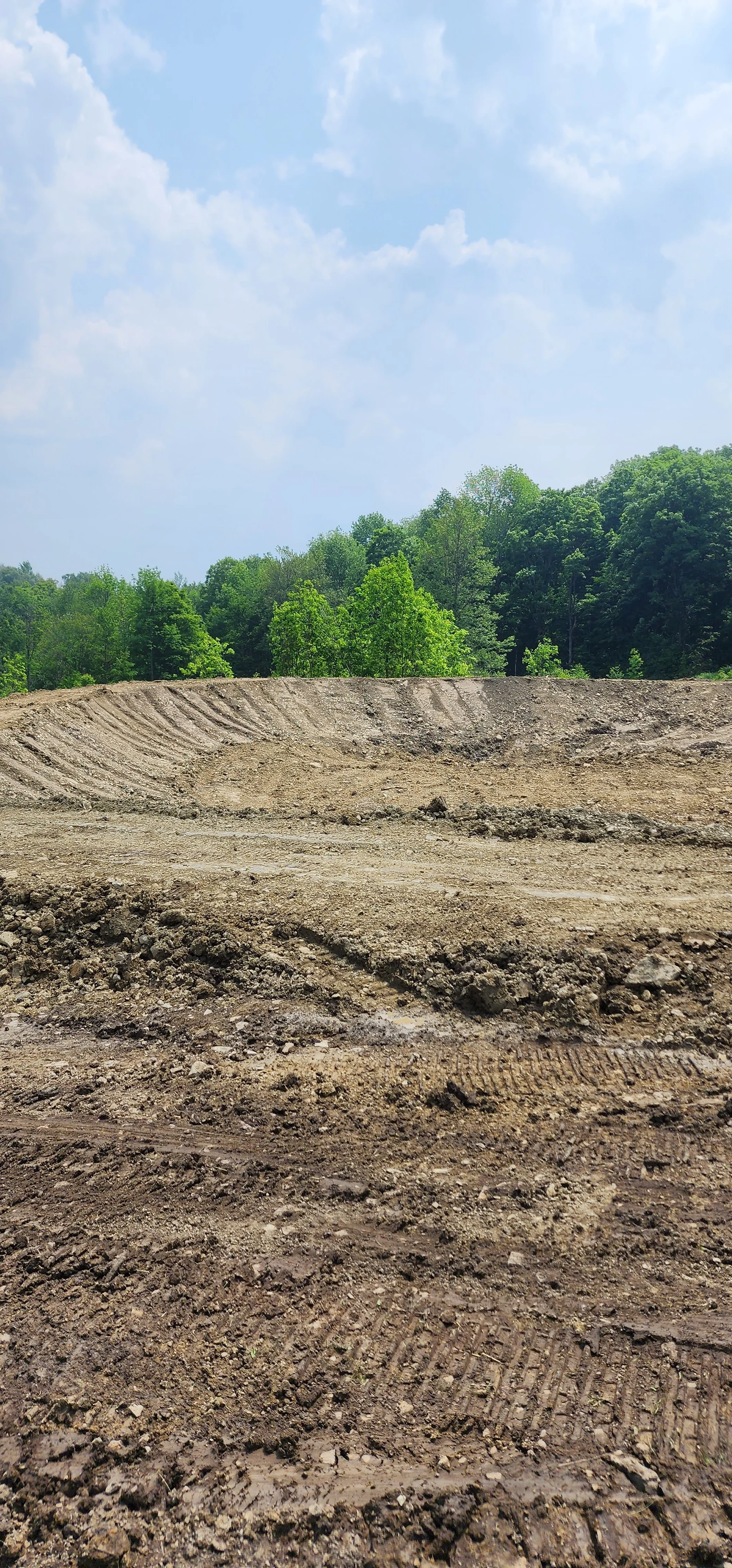 A construction site with freshly dug earth and tire tracks, bordered by green trees under a partly cloudy sky.