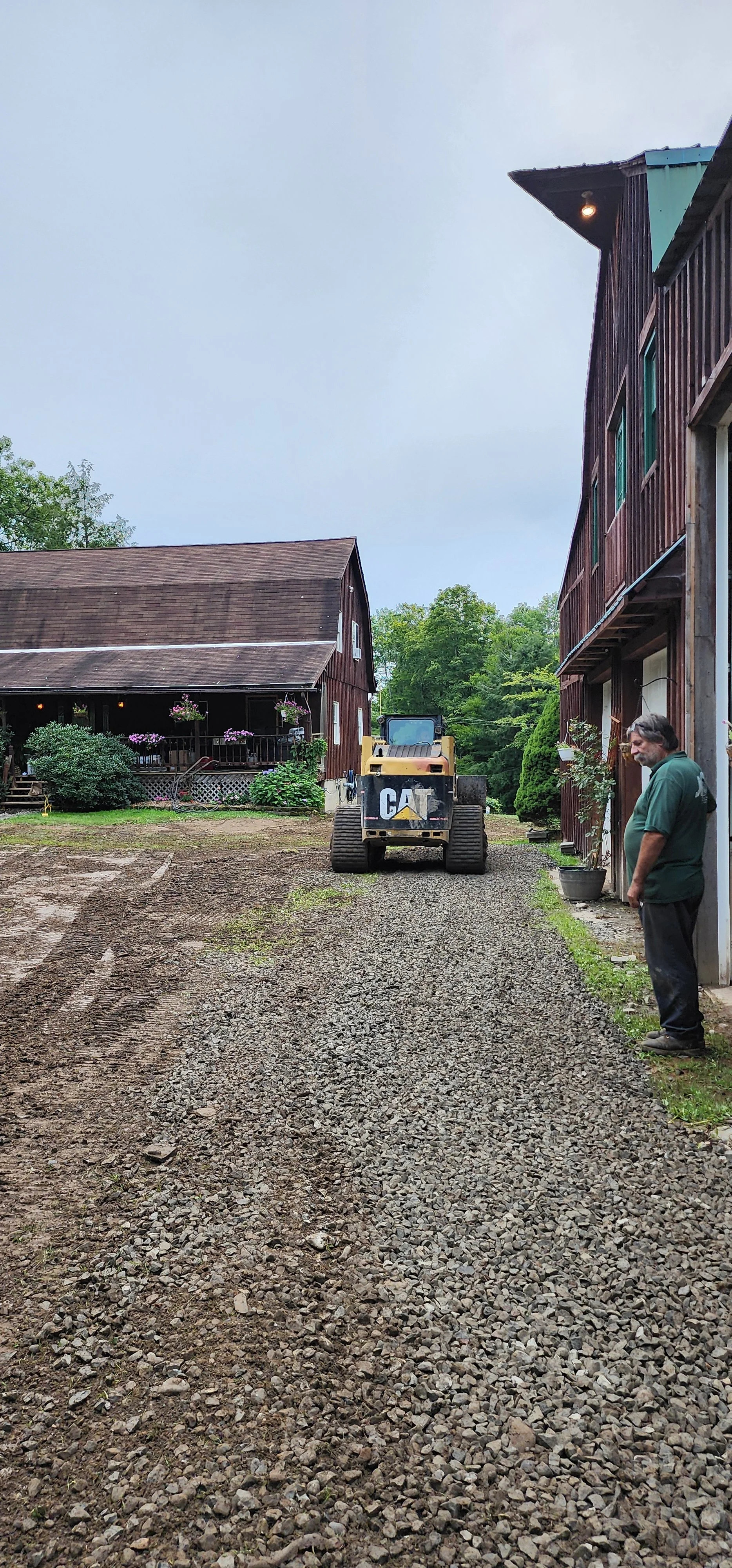 A man standing on a gravel driveway next to a two-story wooden building with a man on the right, a yellow CAT bulldozer in the middle, and another wooden building with a porch and hanging flower baskets on the left, surrounded by green trees.