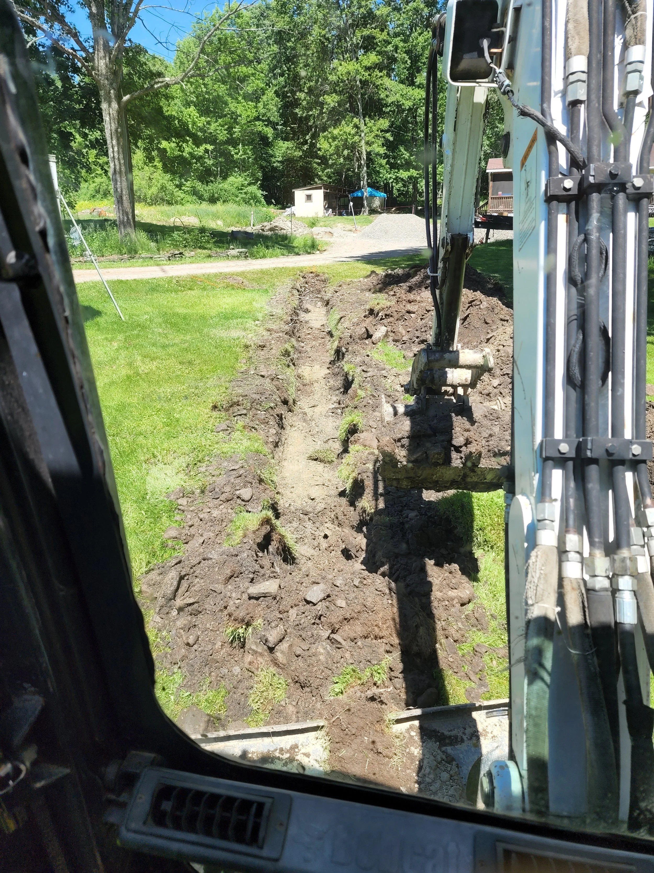 View from inside a construction excavator looking at a dirt digging site with a narrow trench and green grass on either side, trees and a small shed in the background.