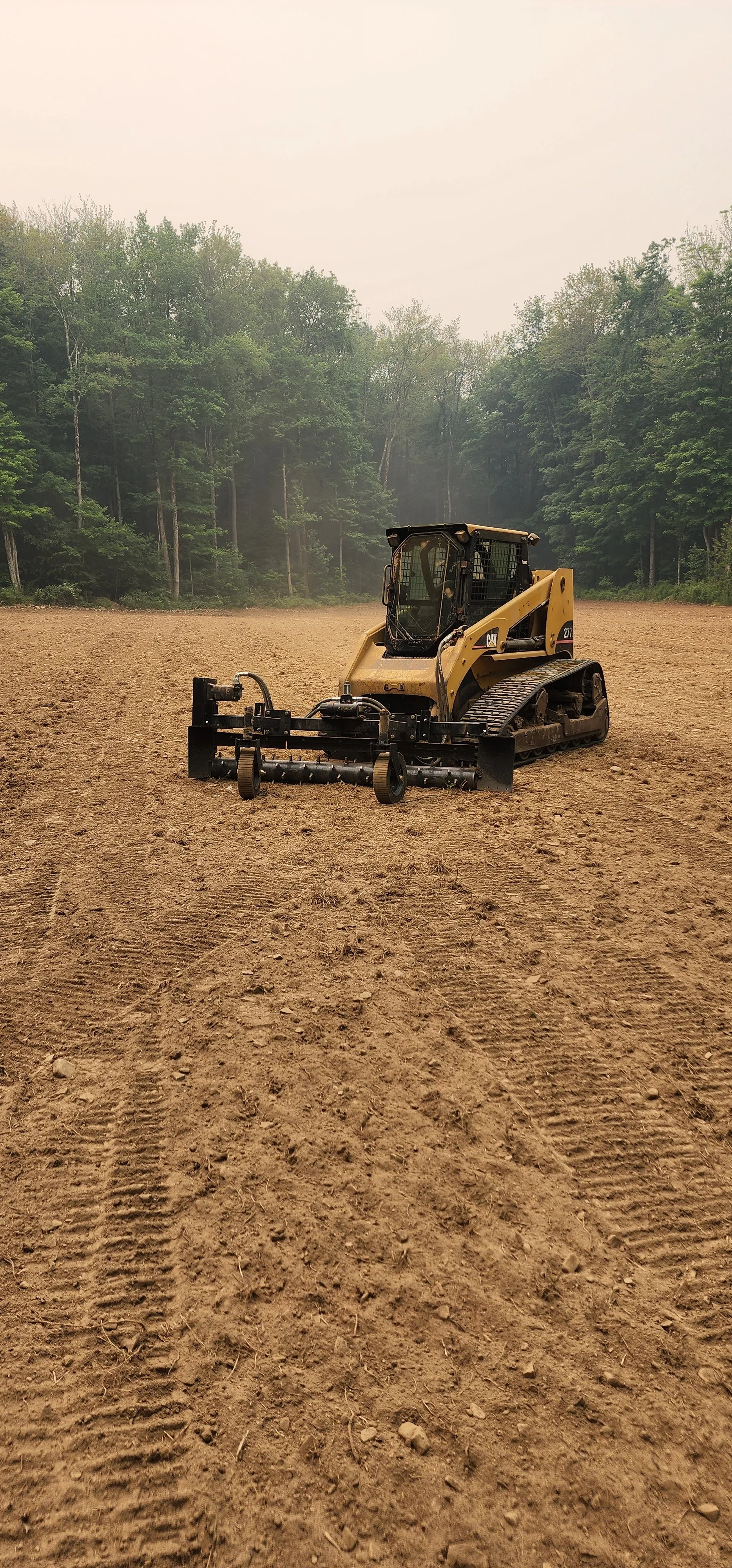 A yellow and black built bulldozer on a large dirt field, with a forest in the background under an overcast sky.