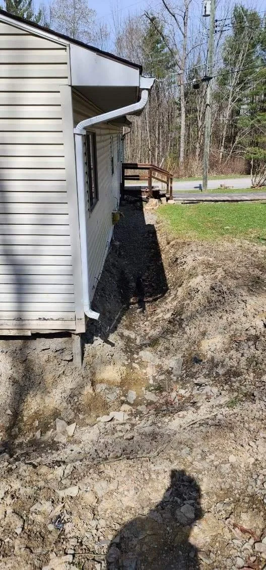 Side view of a house with a drainage pipe extending from the roof, and a damaged yard with exposed dirt and rocks.
