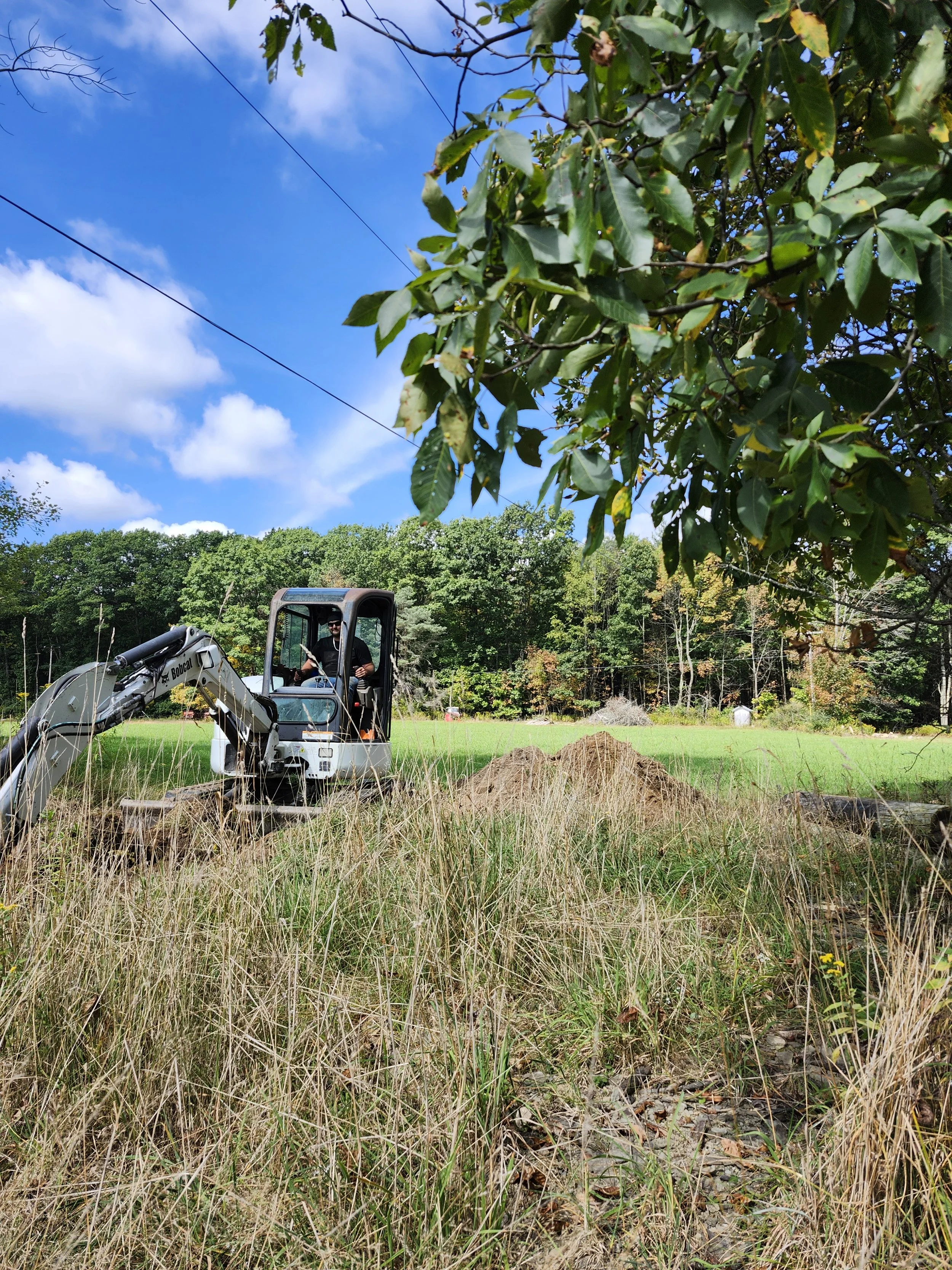 A person operating a mini excavator in a grassy field with trees in the background and a partly cloudy sky.