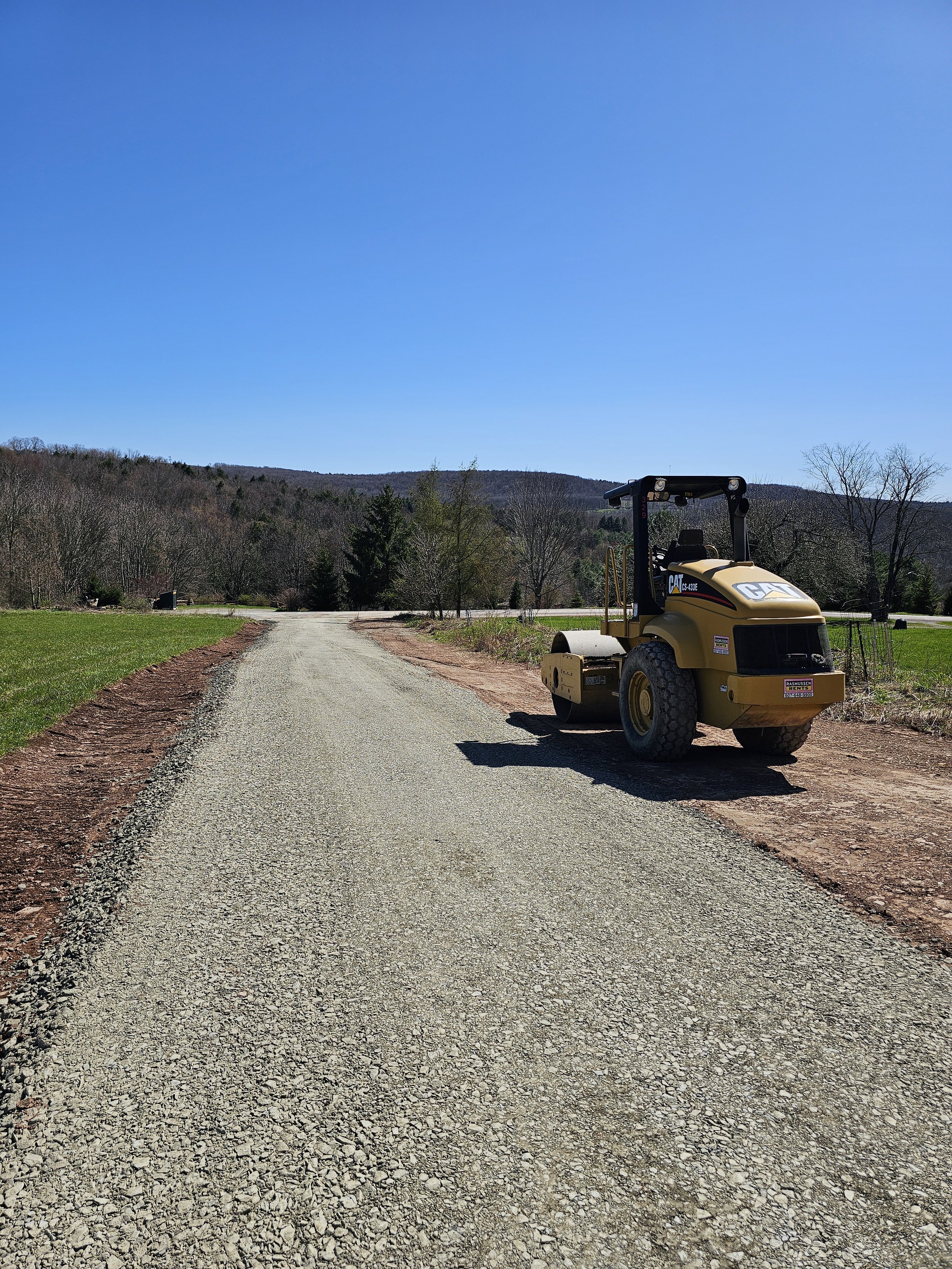 A construction vehicle parked on a gravel path in a rural area with trees and hills in the background under a clear blue sky.