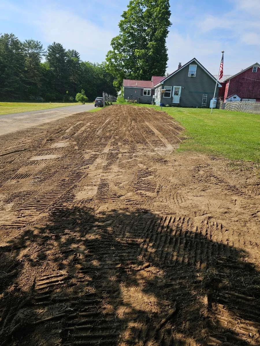 A dirt path with tire tracks in front of a house with a gray exterior, red roof, and American flag, surrounded by green grass and trees under a blue sky.