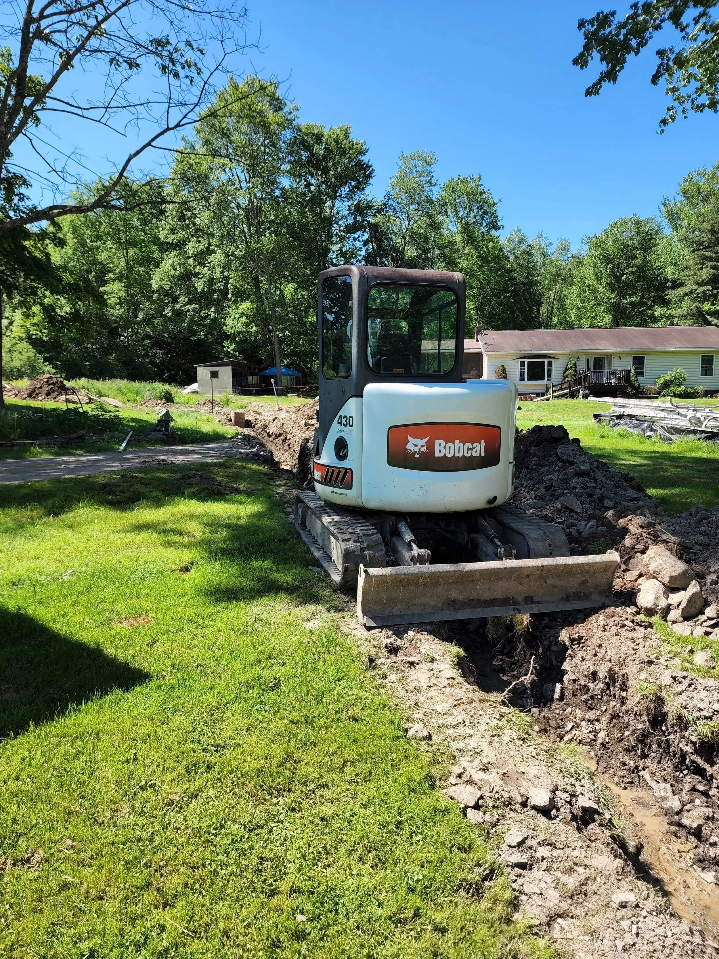 A small Bobcat excavator digging a trench in the lawn of a residential yard on a sunny day.