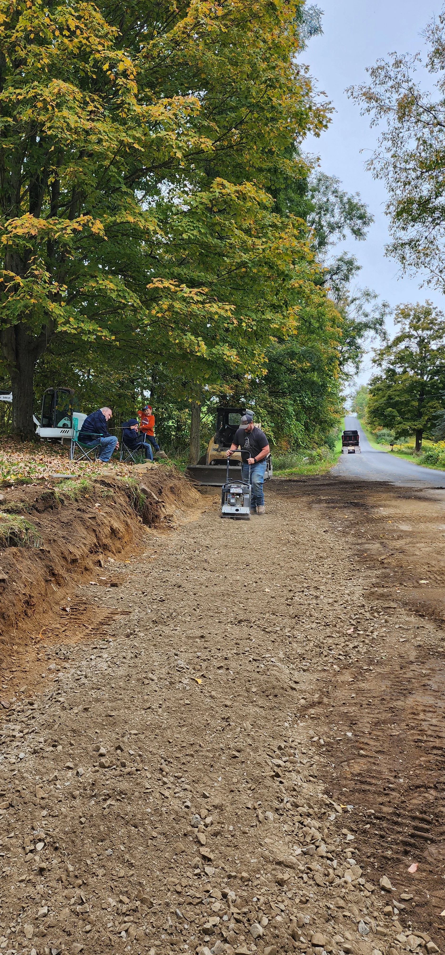 Workers leveling a dirt road with a compacting machine while several people sit and chat under trees with green and yellow fall leaves.
