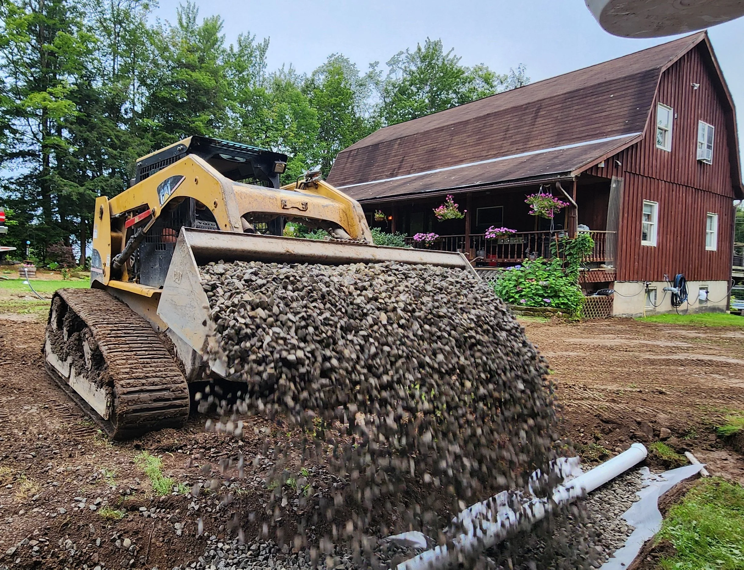 A small bulldozer spreading gravel on a construction site in front of a red wooden house with flowering plants on the porch.