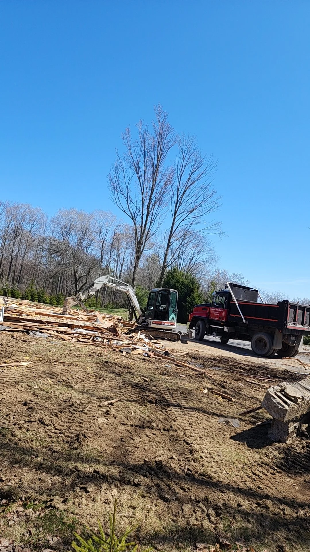 Construction site with a small excavator and a truck, piles of wood debris, bare trees, and clear blue sky.