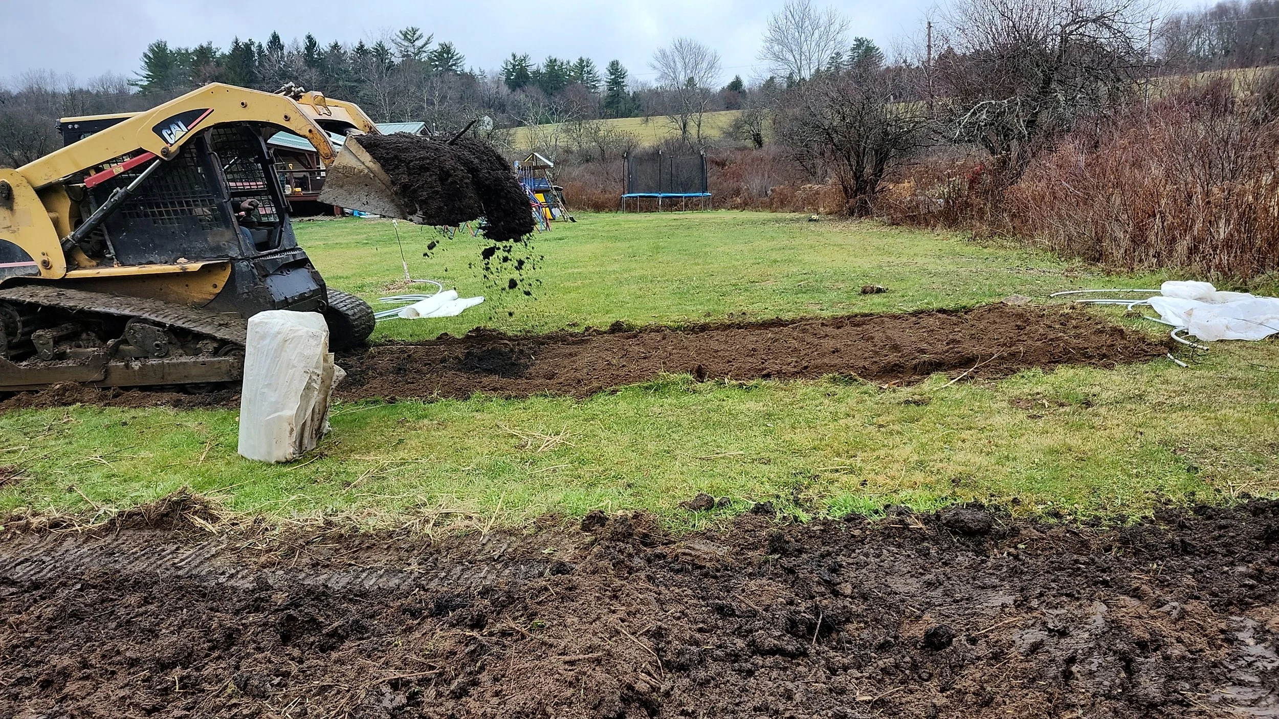 A small excavator digging a trench in a grassy yard with trees, a trampoline, and a shed in the background