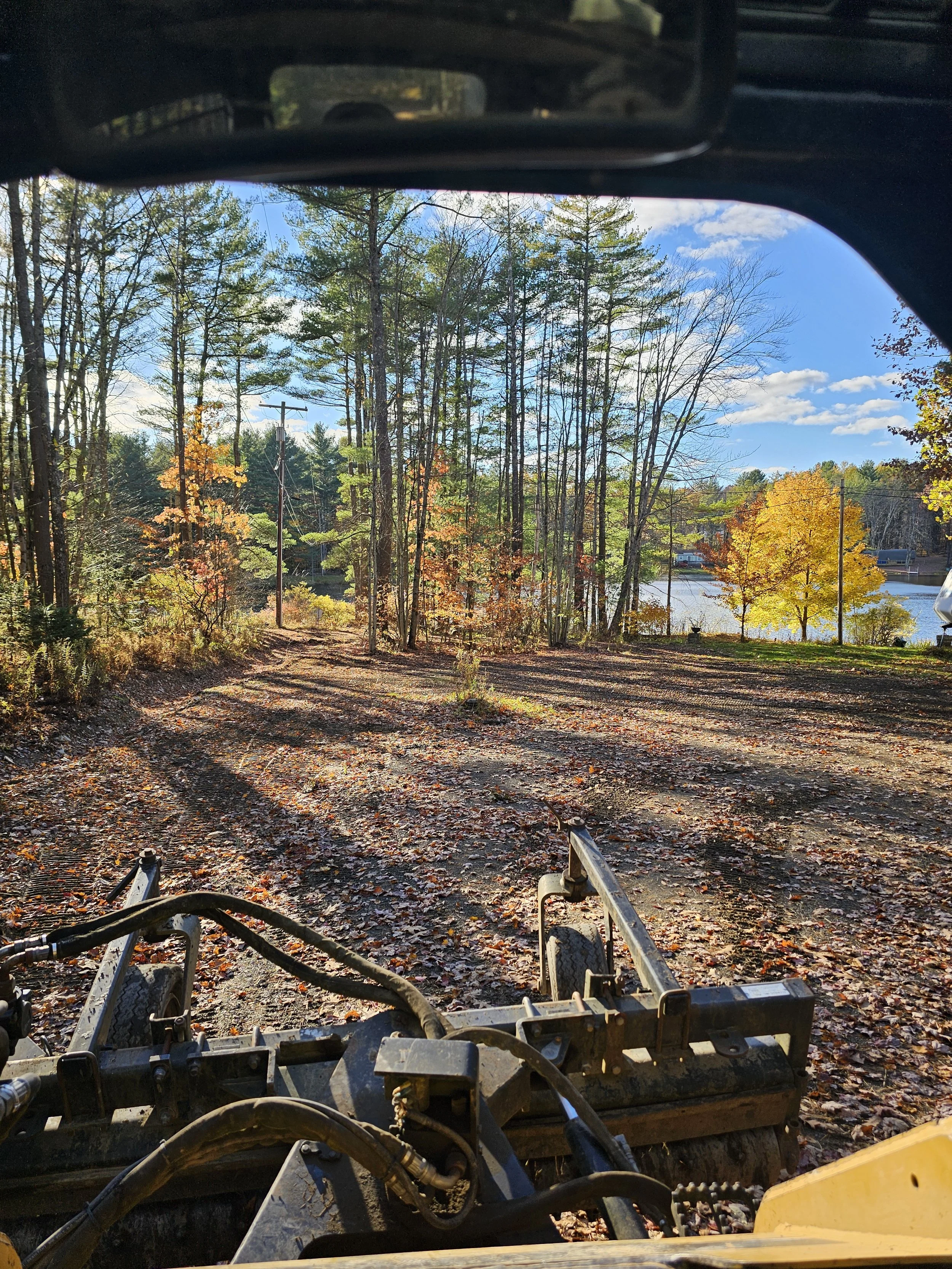 View of a yard with leaves on the ground and a rake attachment inside a vehicle, with trees, a lake, and a blue sky in the background, seen through the windshield.
