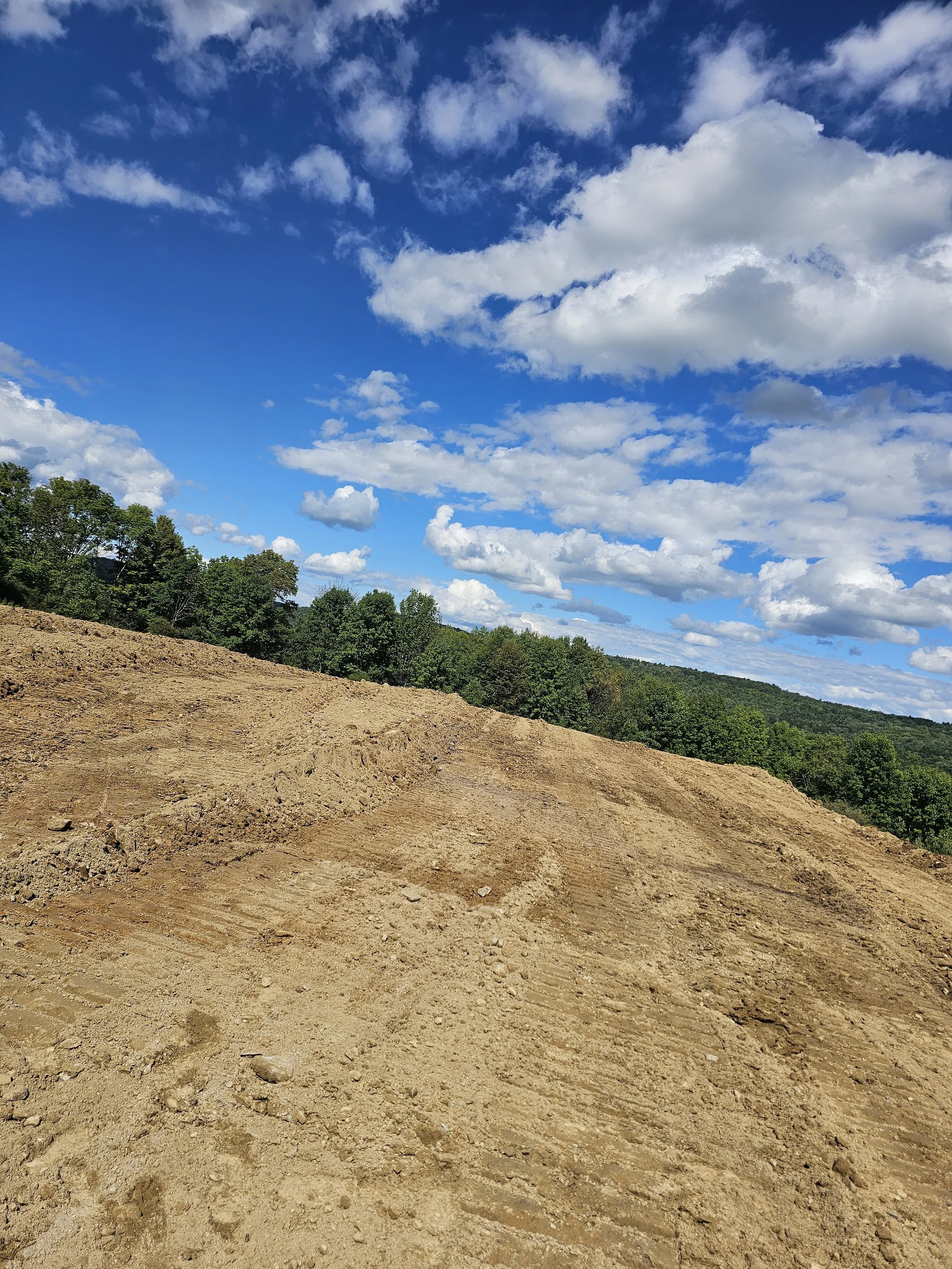 A landscape view featuring a dirt hill with tire tracks in the foreground, a green treeline in the middle distance, and a bright blue sky with scattered white clouds overhead.